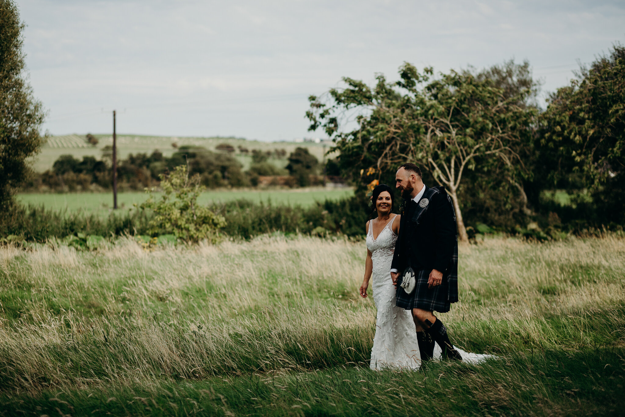 bride wearing unbridaled dress walking with groom in field outdoors at dalduff farm at their barn wedding photographed by wedding photographer ayrshire scotland