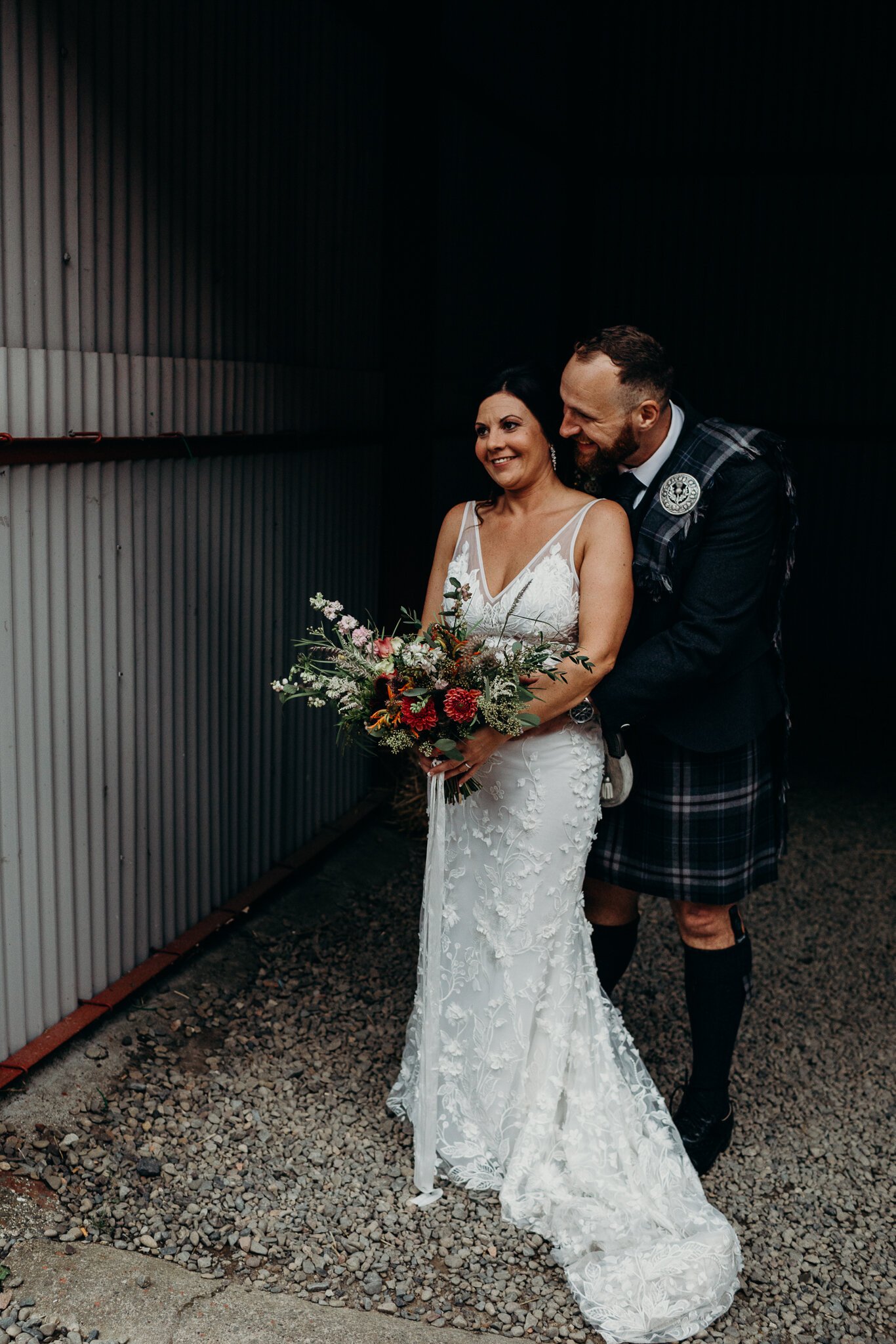 groom hugging bride wearing unbridaled dress holding bouquet by sparrow &amp; rose flowers in barn at dalduff farm photographed by wedding photographer ayrshire Scotland