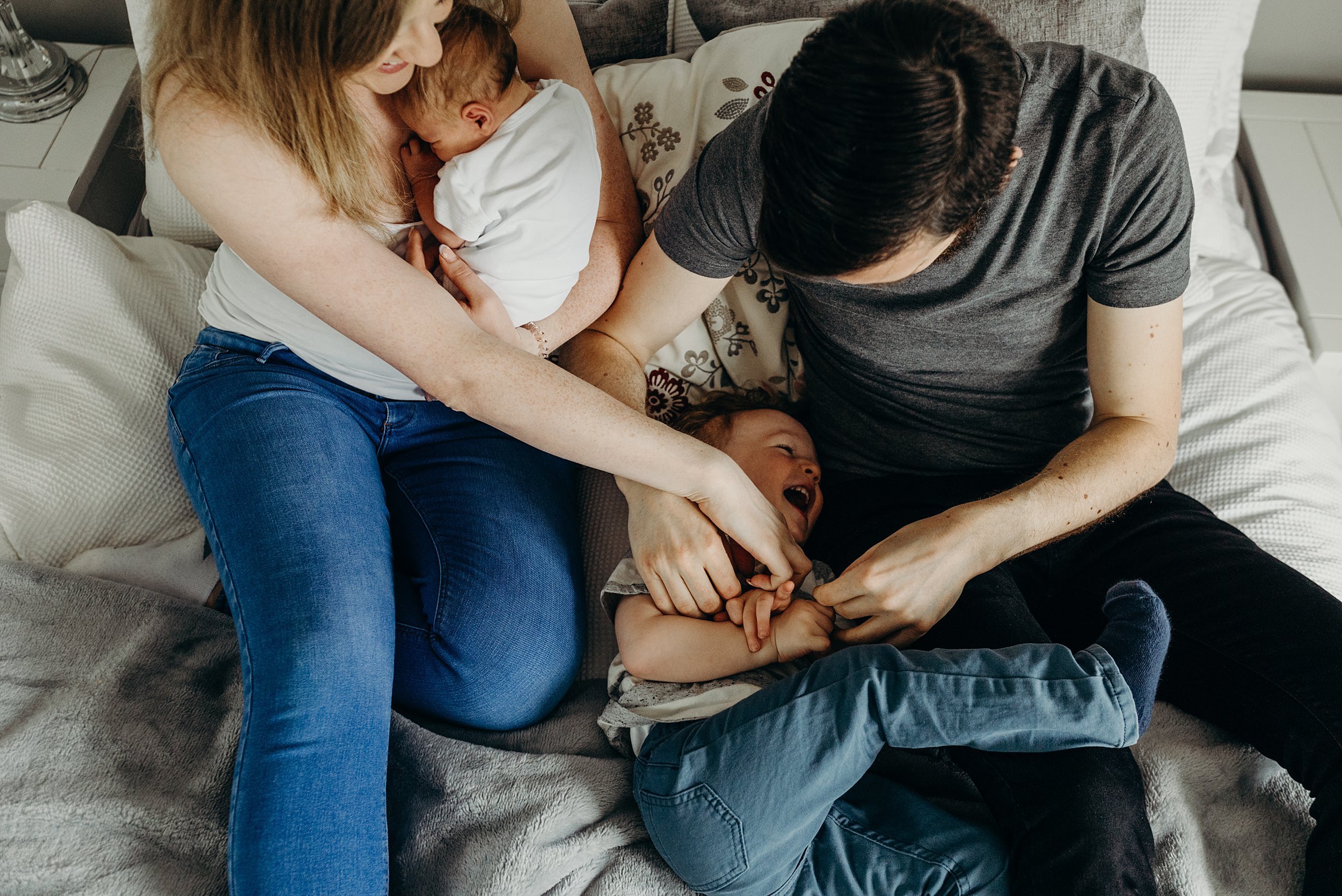 mum and dad smile and hug toddler and baby on bed during photo session baby photography glasgow