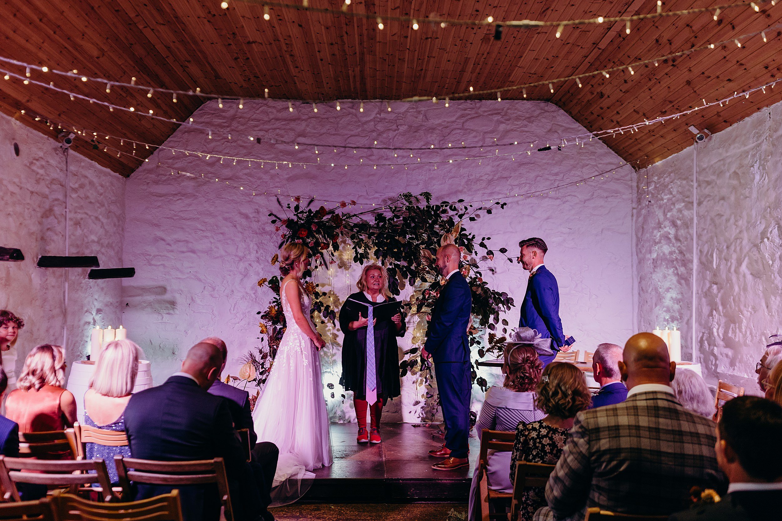 bride and groom with humanist celebrant during their wedding ceremony at dalduff farm with fairy lights and flower arch shown in dalduff farm wedding photos