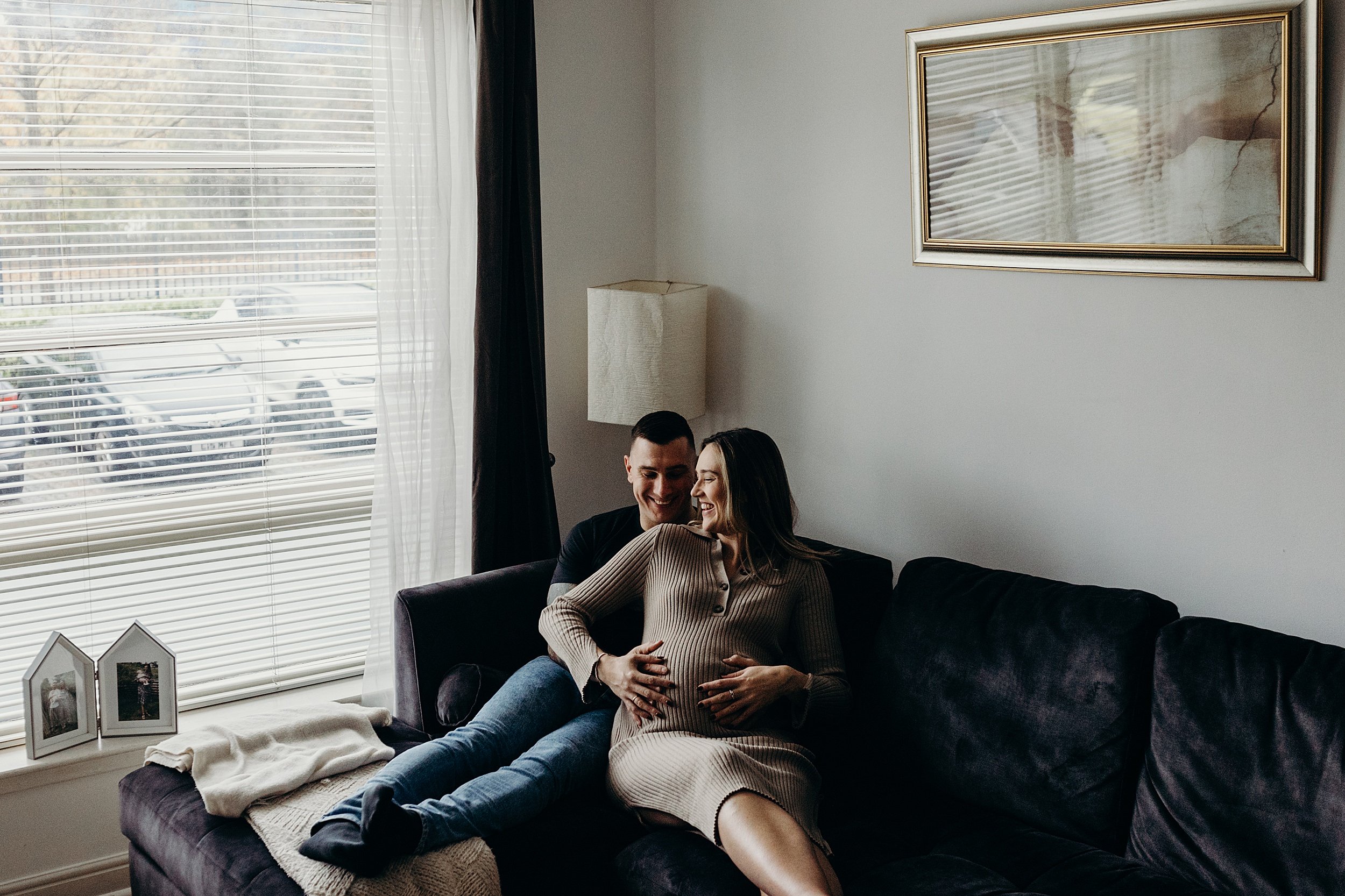 couple sitting cuddling on their sofa beside a window during a photo session by a maternity photographer glasgow