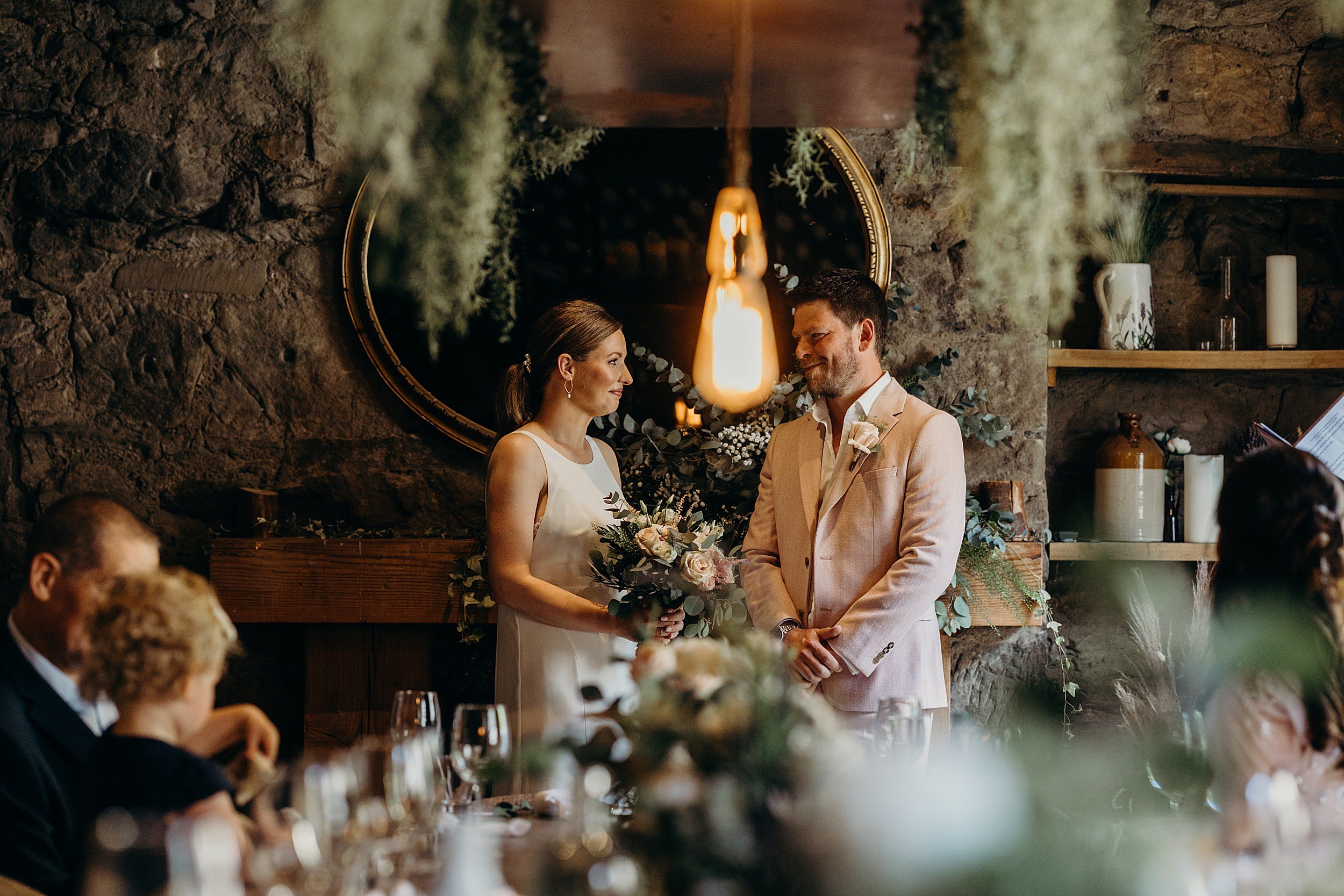 bride and groom looking at each other and smiling during ceremony with hanging green foliage and pendant lights at the bothy glasgow wedding