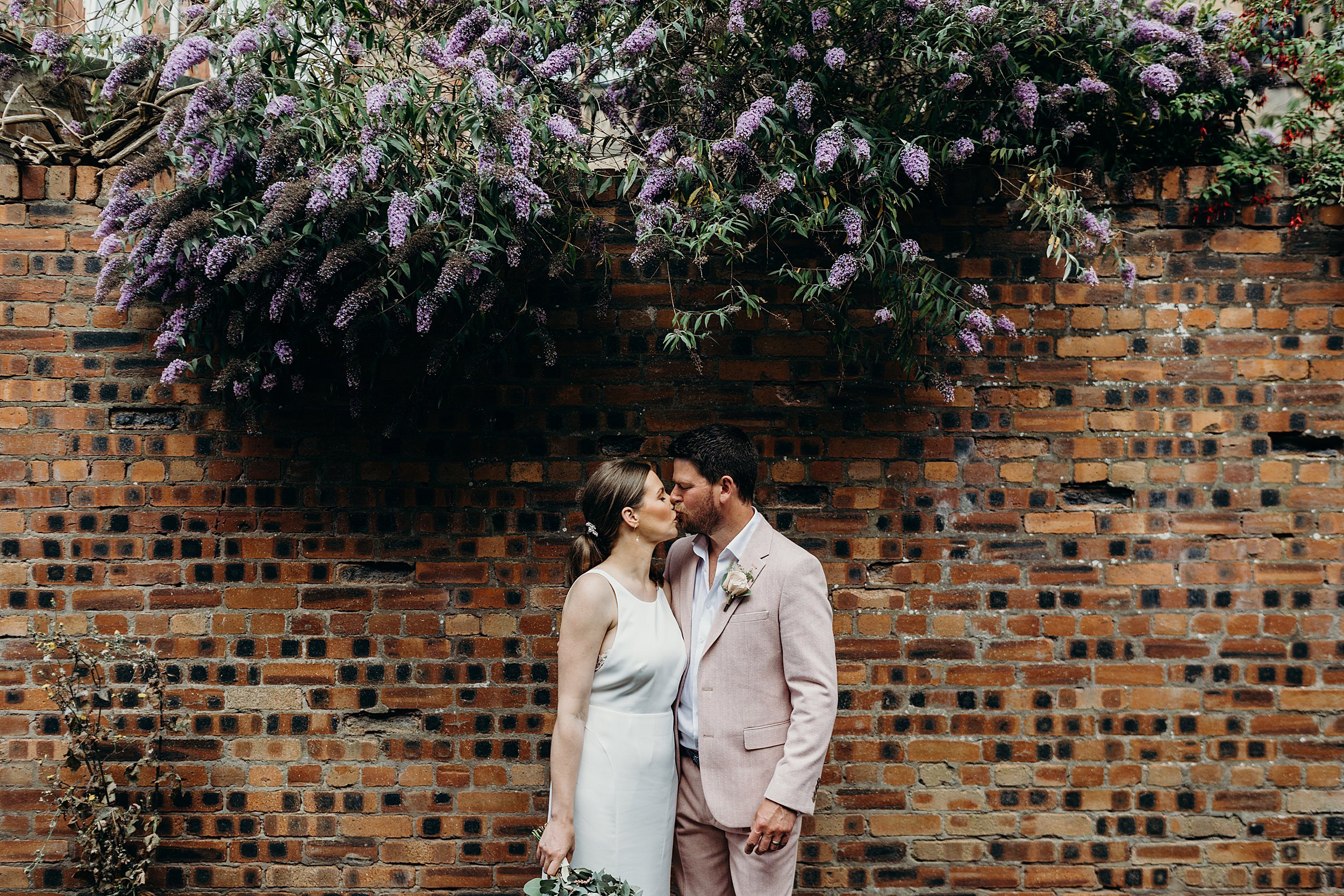bride and groom kissing in front of brick wall underneath purple flowers at their mini wedding in the west end glasgow