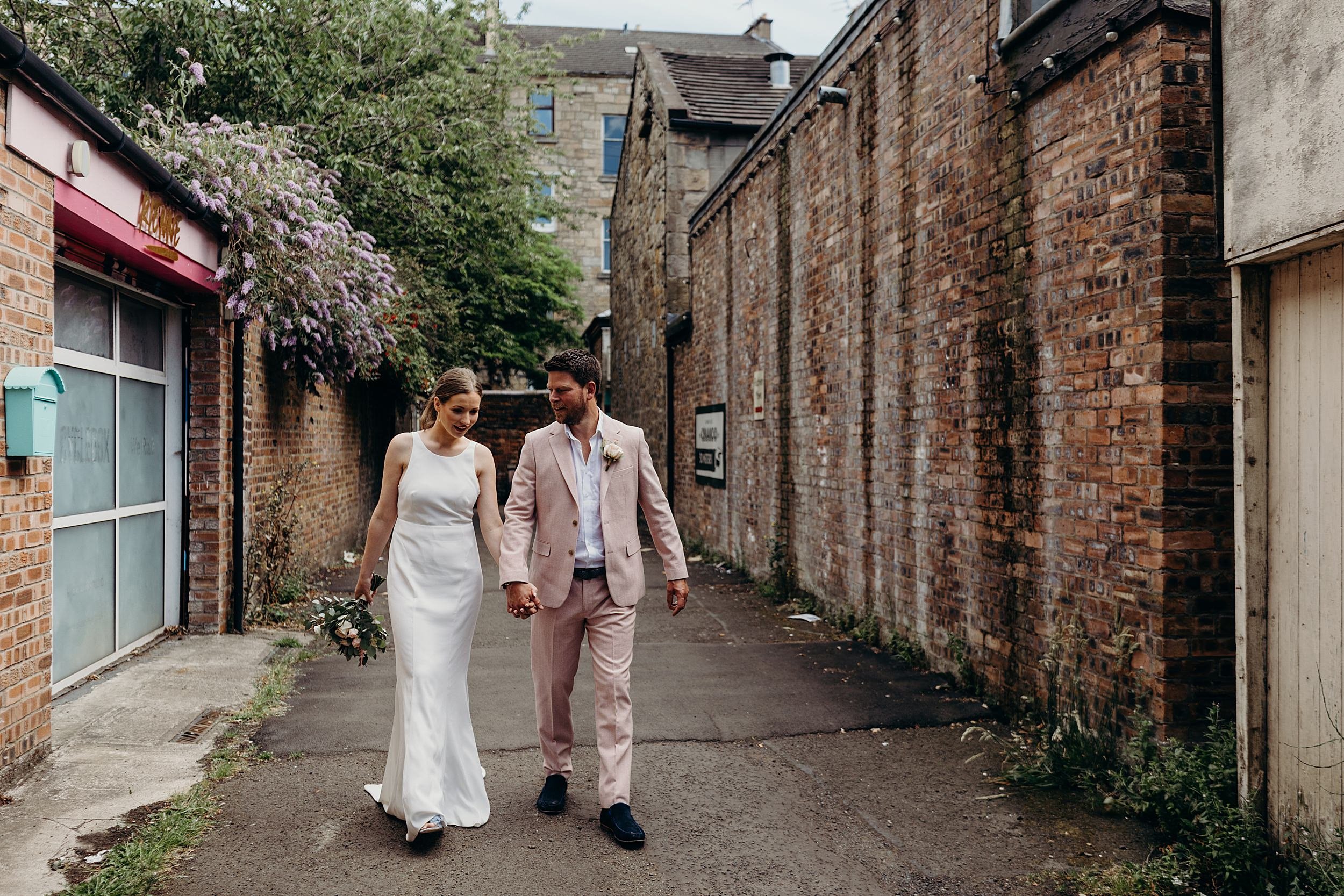 bride and groom in pink suit walking down a glasgow lane after their ceremony at the bothy glasgow wedding