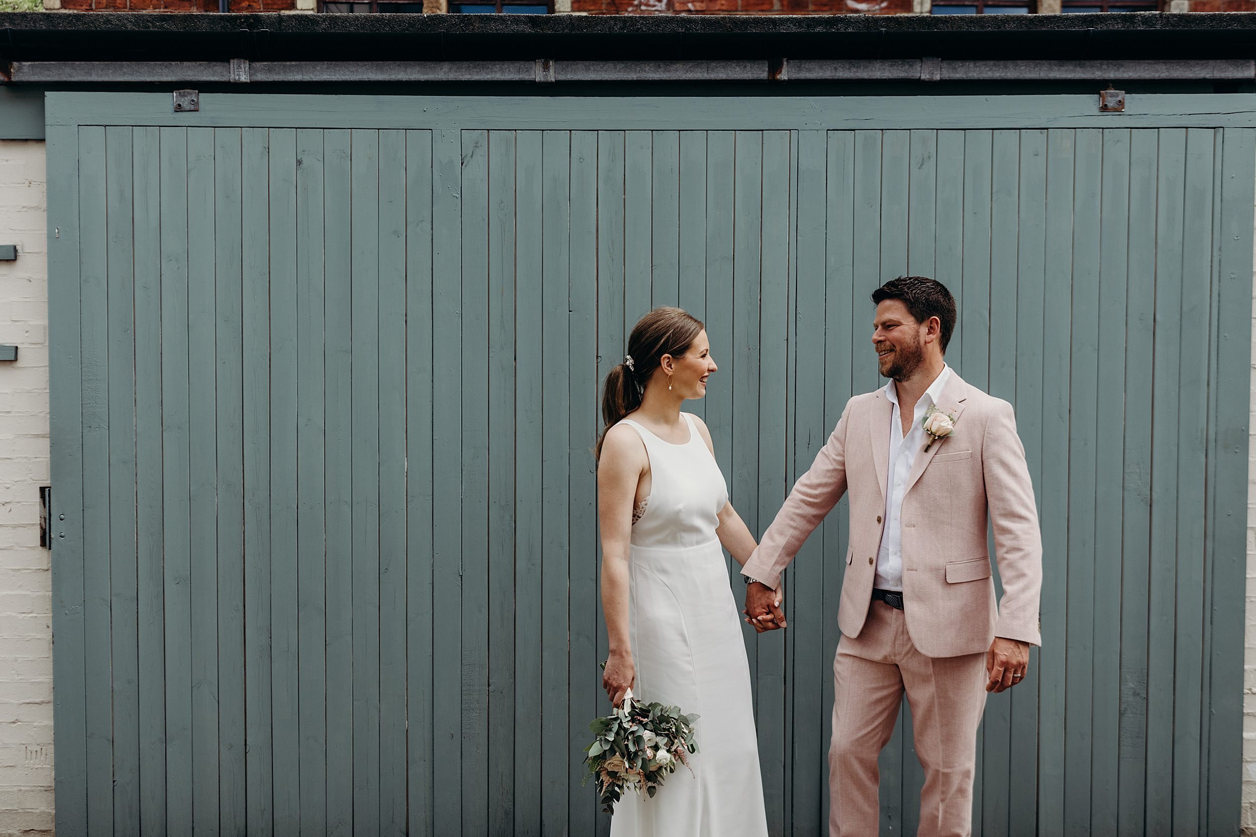 bride and groom looking at each other and smiling in front of pale green wooden wall after their ceremony at the bothy glasgow wedding