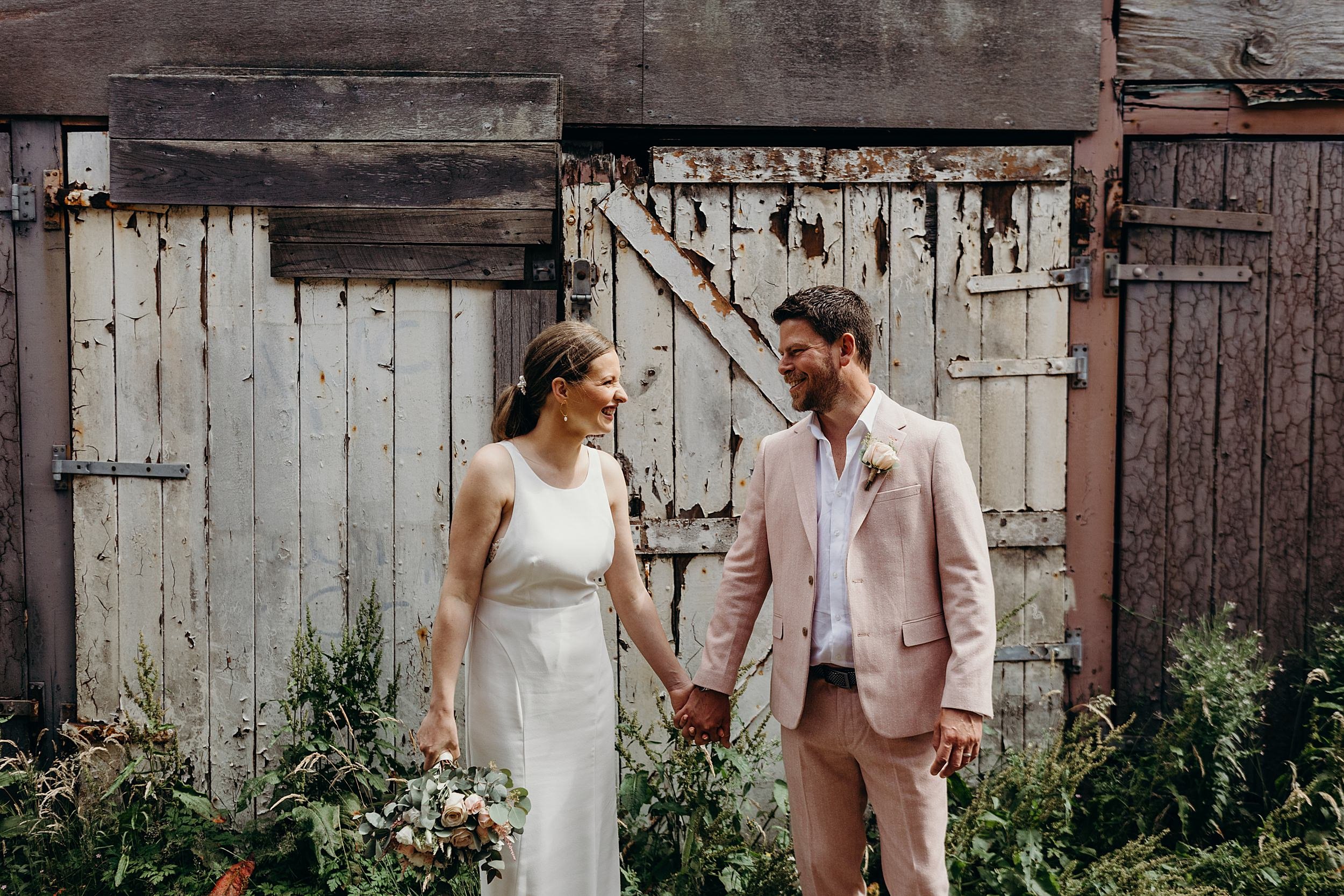 bride and groom holding hands and smiling at each other while standing in front of rustic old doors after their ceremony at the bothy glasgow wedding