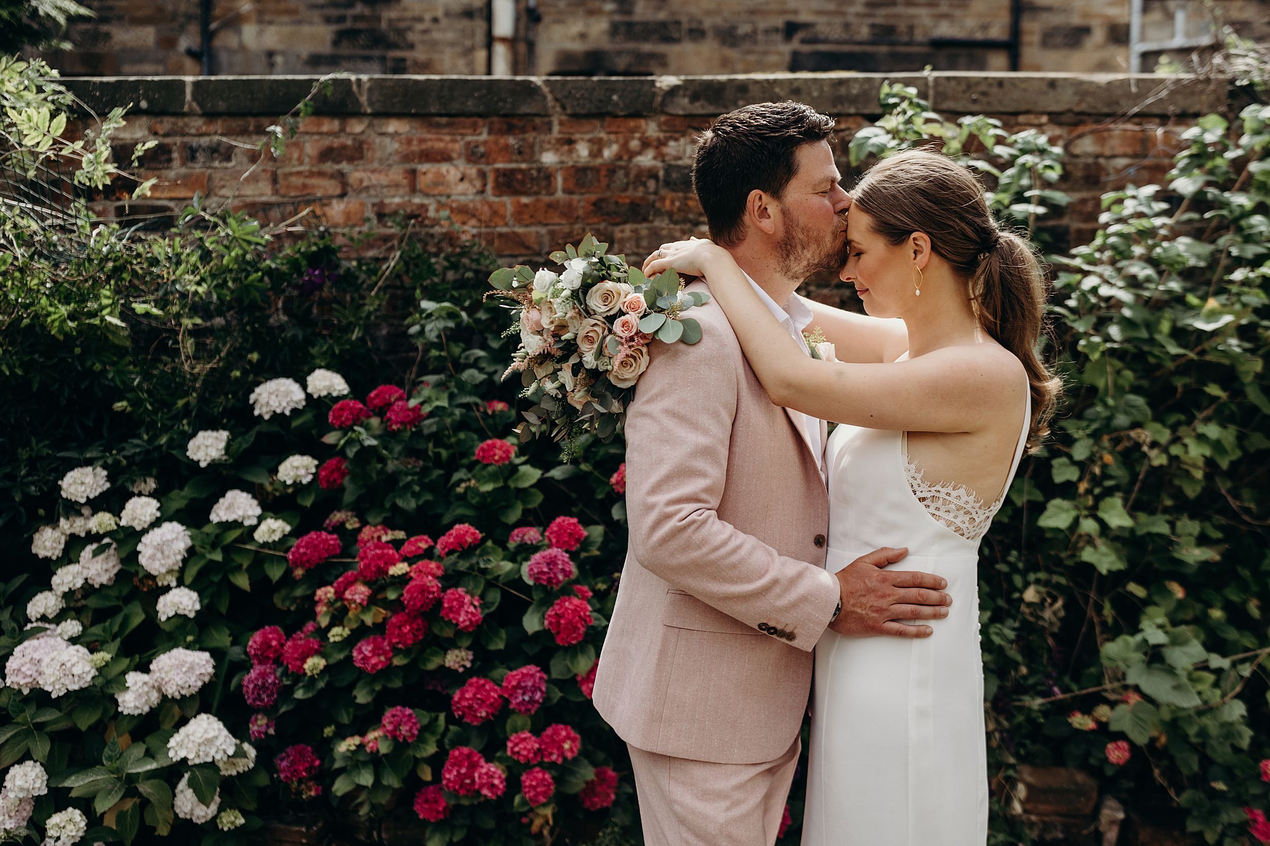 groom kissing brides forehead surrounded by flowers and rustic brick wall after micro wedding in the west end glasgow