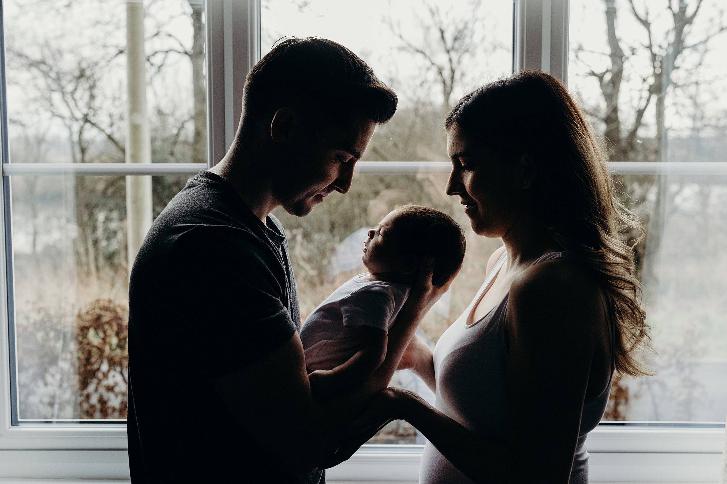newborn photography glasgow shot of a father and mother standing in front of a window holding their baby boy