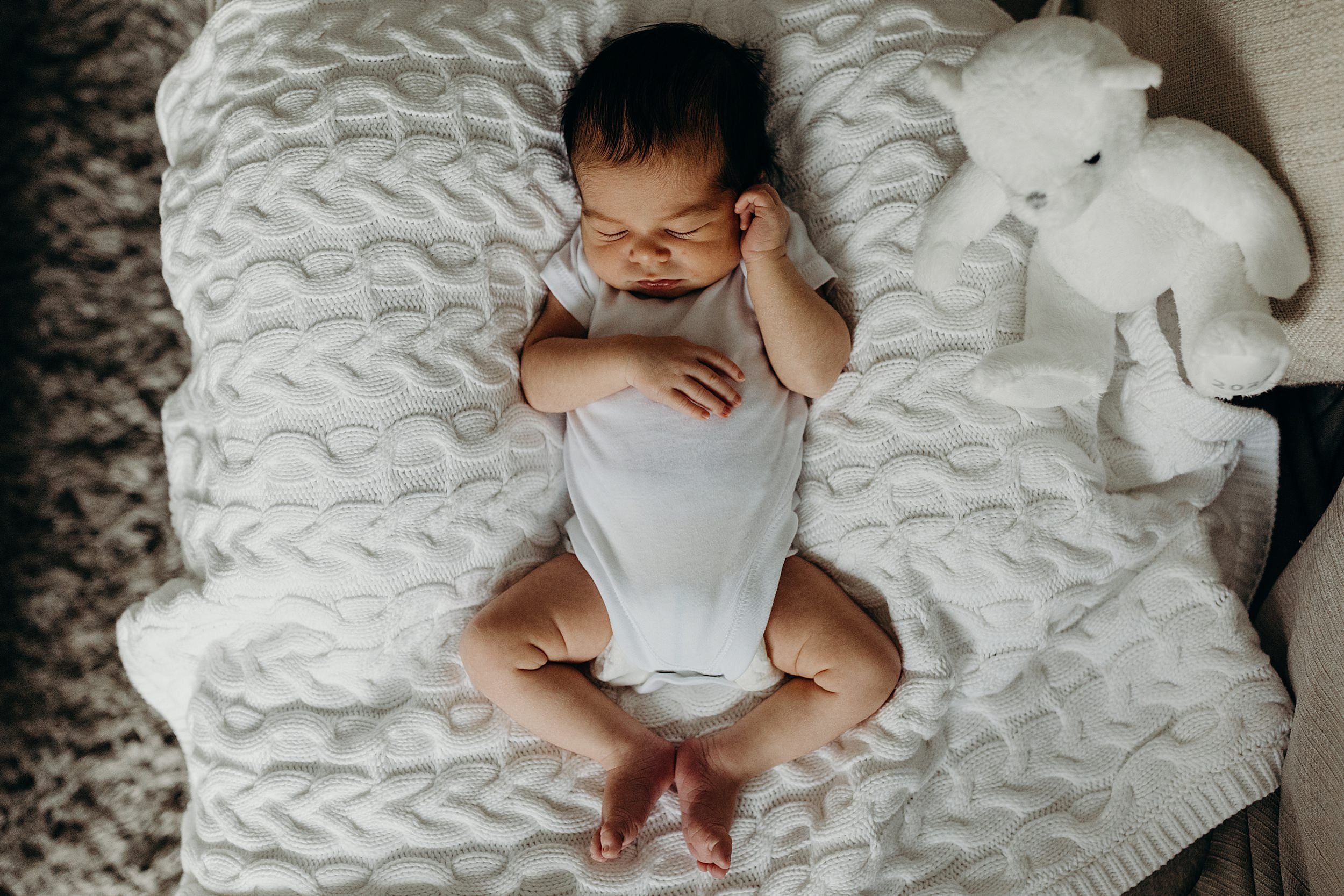 baby boy in a white babygrow lying on a white blanket beside a white teddy bear during newborn photography glasgow scotland shoot