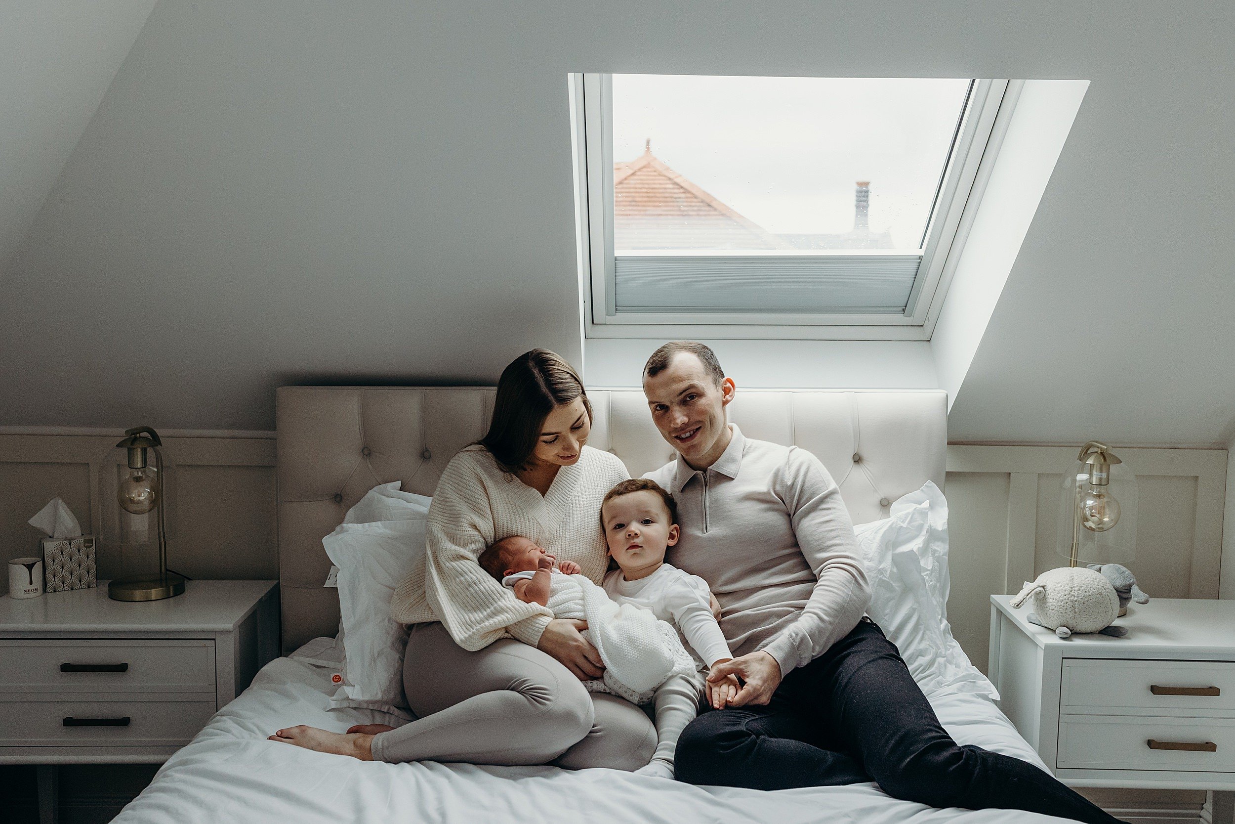 family photographer glasgow captures mother father toddler boy and baby girl sitting on a double bed in the parents bedroom