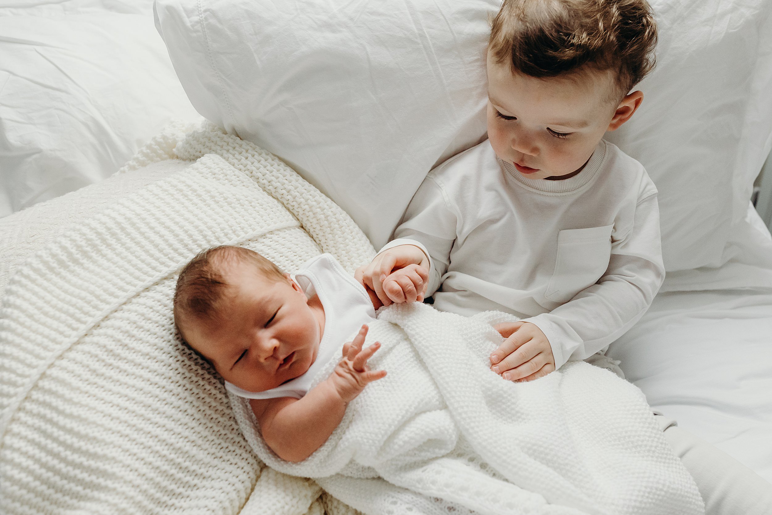 family photographer glasgow scotland captures toddler boy and baby on white blanket and pillows the toddler is holding the baby's hand
