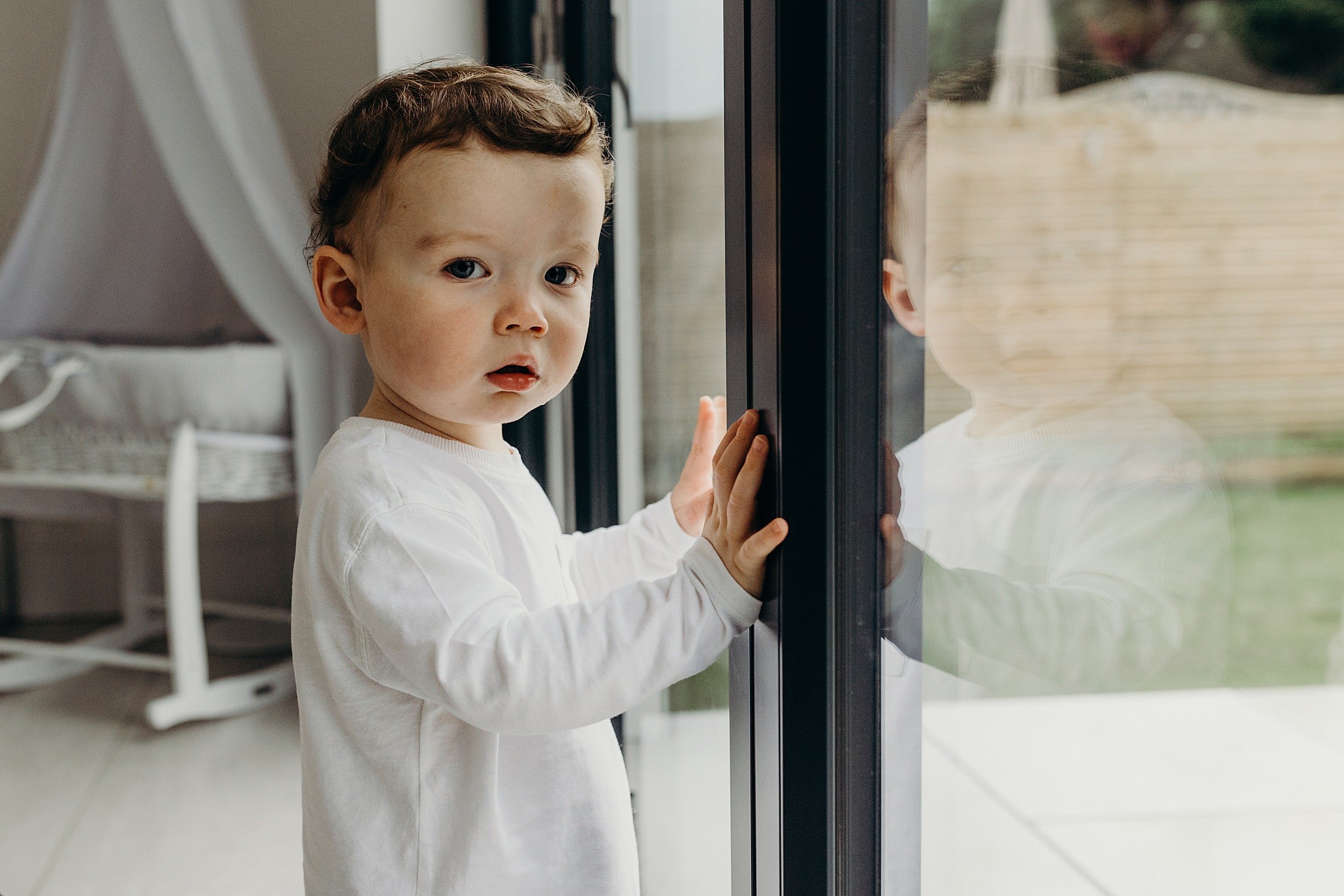 family photographer glasgow scotland captures toddler boy with red hair standing by patio doors with crib in the background