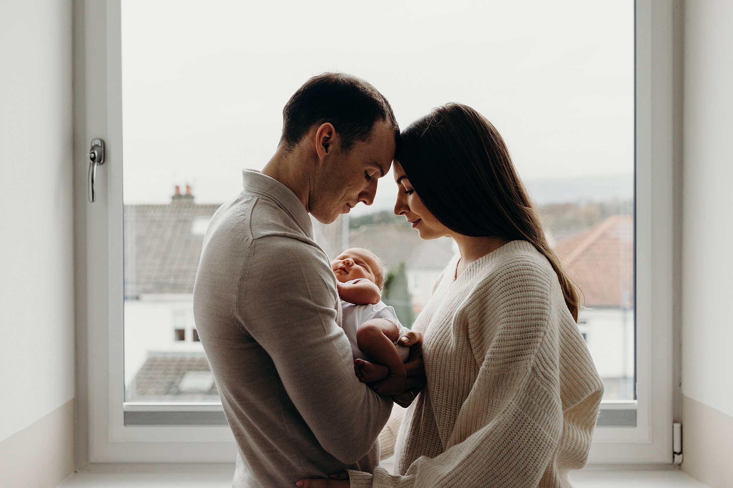 family photographer glasgow captures father holding baby he is forehead to forehead with mother in front of a window with houses visible outside