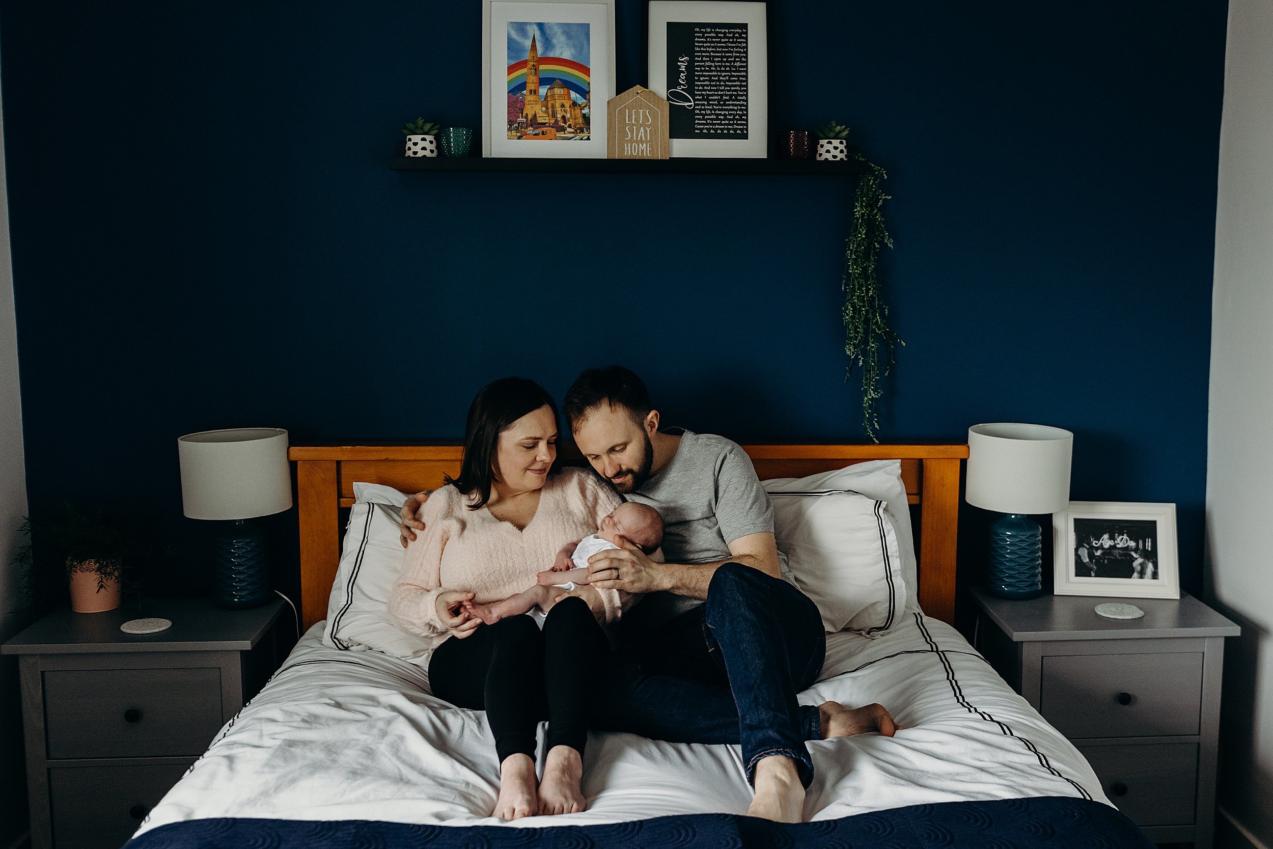 mother and father sit on their double bed together holding their baby girl between them during newborn photoshoot glasgow scotland