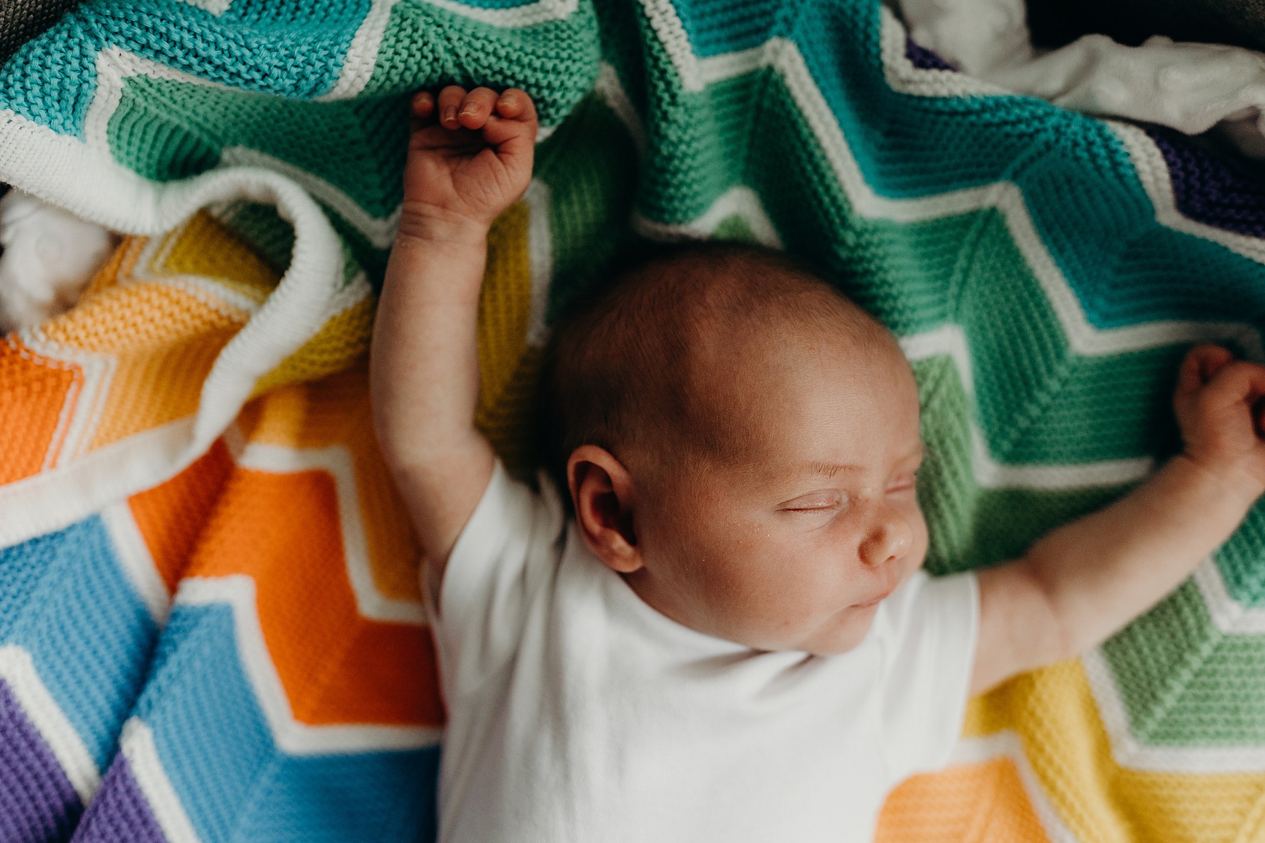 a baby girl lies with eyes closed and arms stretched above her head on a multi coloured knitted blanket during newborn photoshoot glasgow scotland