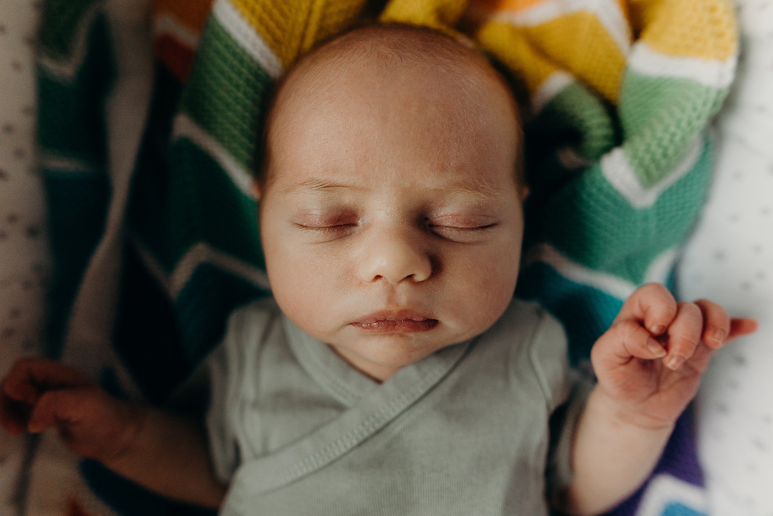 a baby girl lies with eyes closed on a multi coloured knitted blanket during newborn photoshoot glasgow scotland