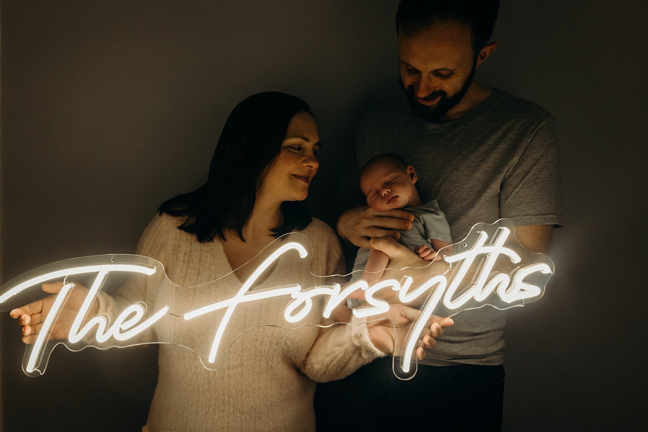 mother smiles and holds a neon sign while father stands next to her holding their sleeping baby girl during newborn photoshoot glasgow scotland