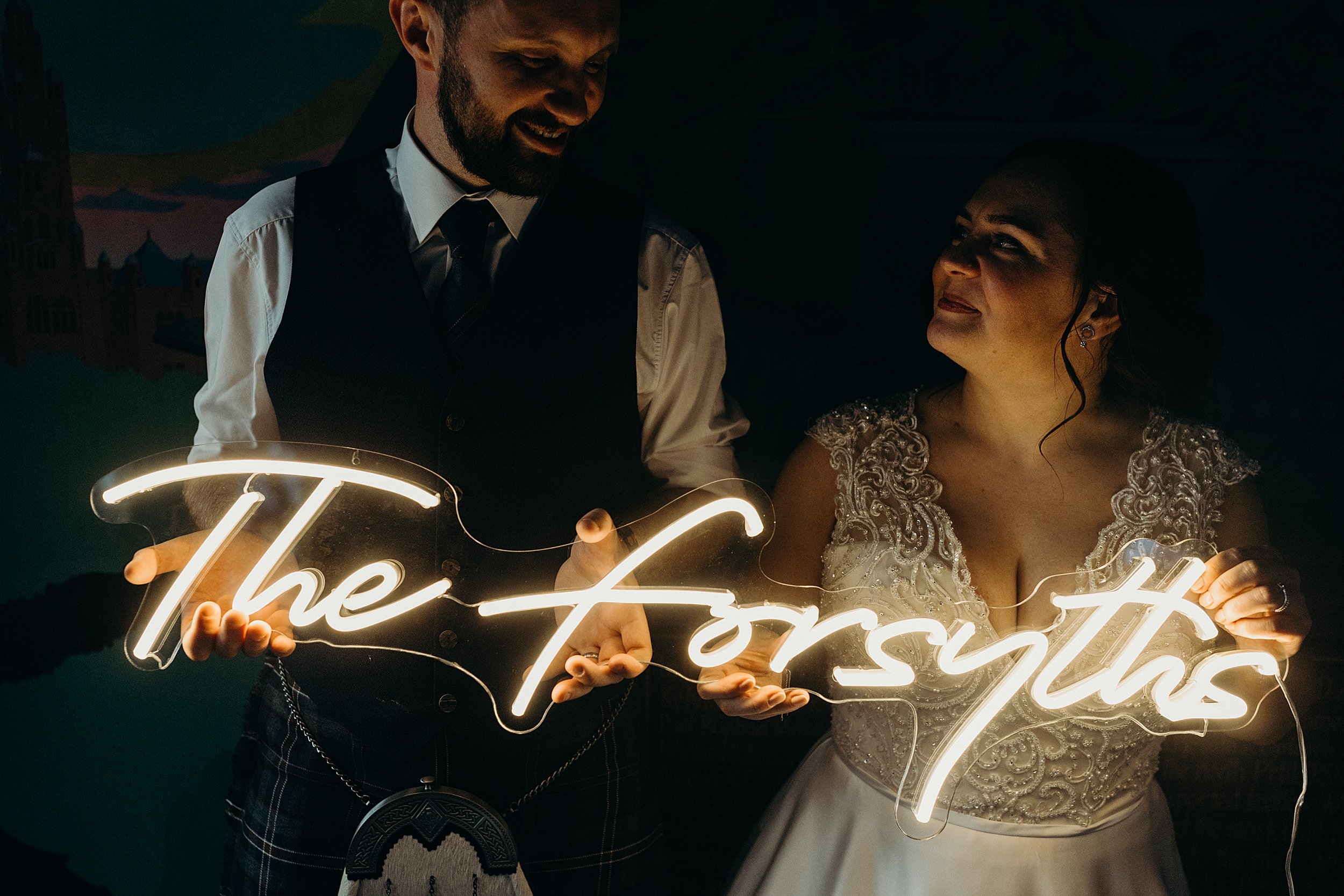 bride and groom stand side by side looking towards each other and smiling they are holding a neon sign