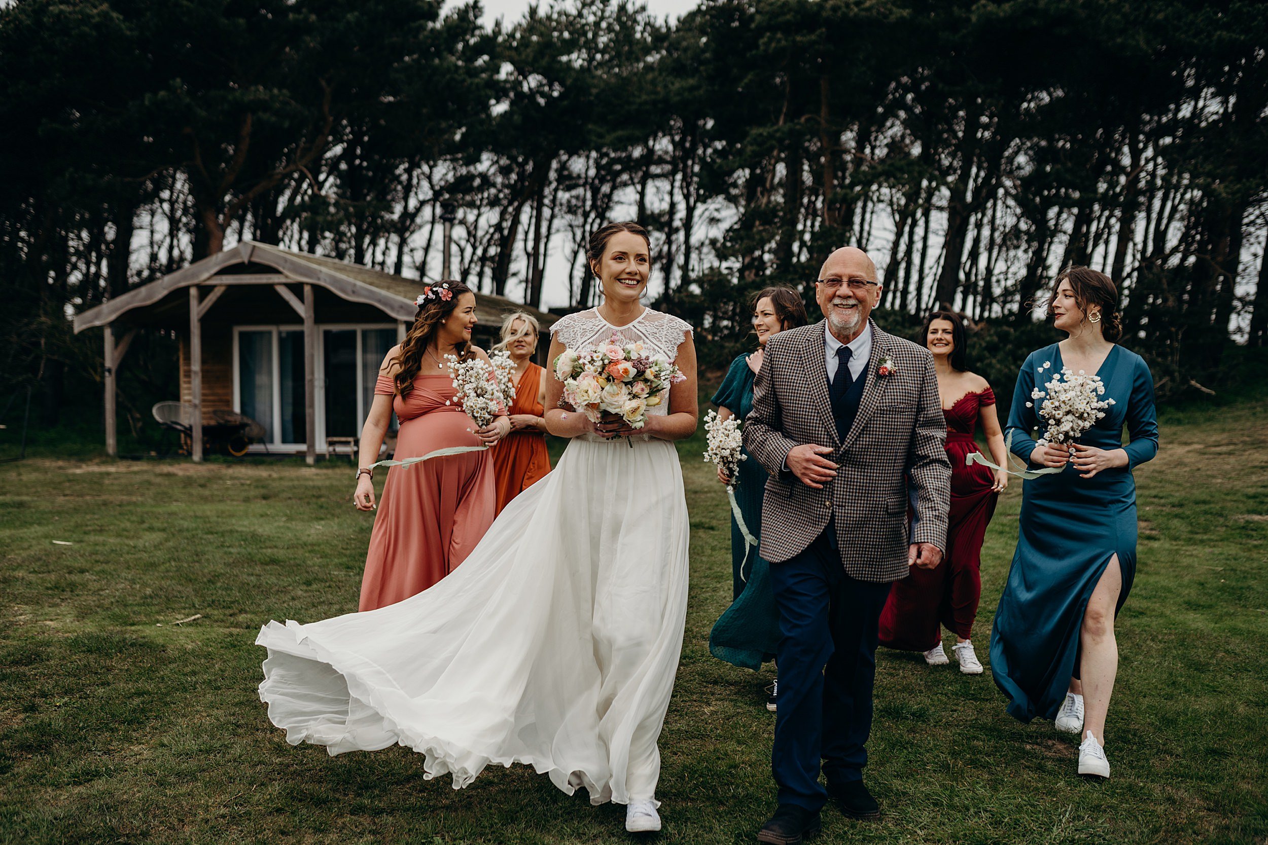a bride and her father arrive for her harvest moon wedding ceremony accompanied by her bridesmaids in colourful dresses trees are visible in the background