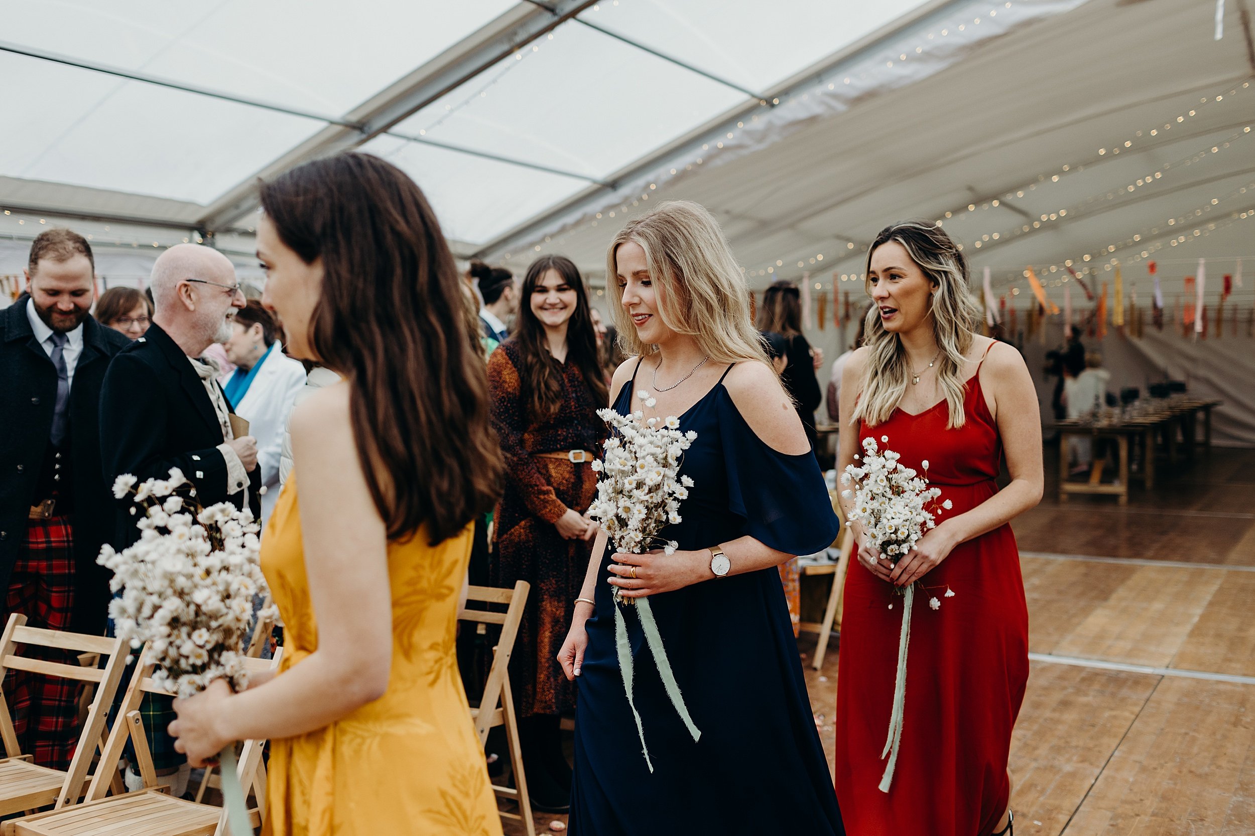 guests look on as three bridesmaids wearing colourful dresses and carrying white bouquets walk down the aisle before a harvest moon wedding ceremony