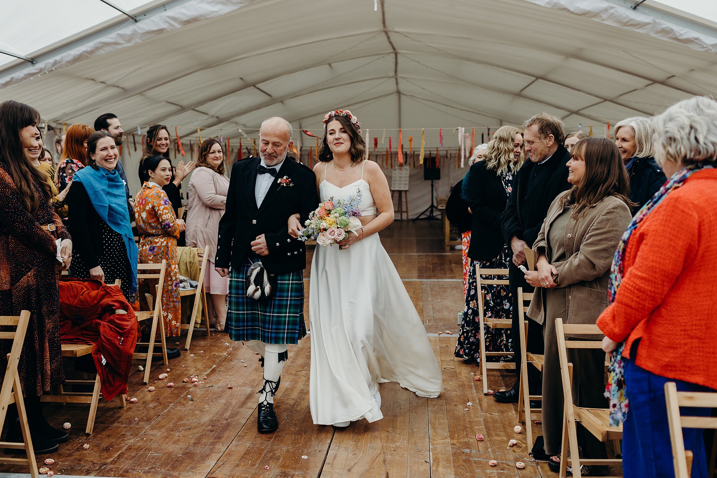 guests watch as a bride walks down the aisle with her father before her harvest moon wedding ceremony inside a white marquee