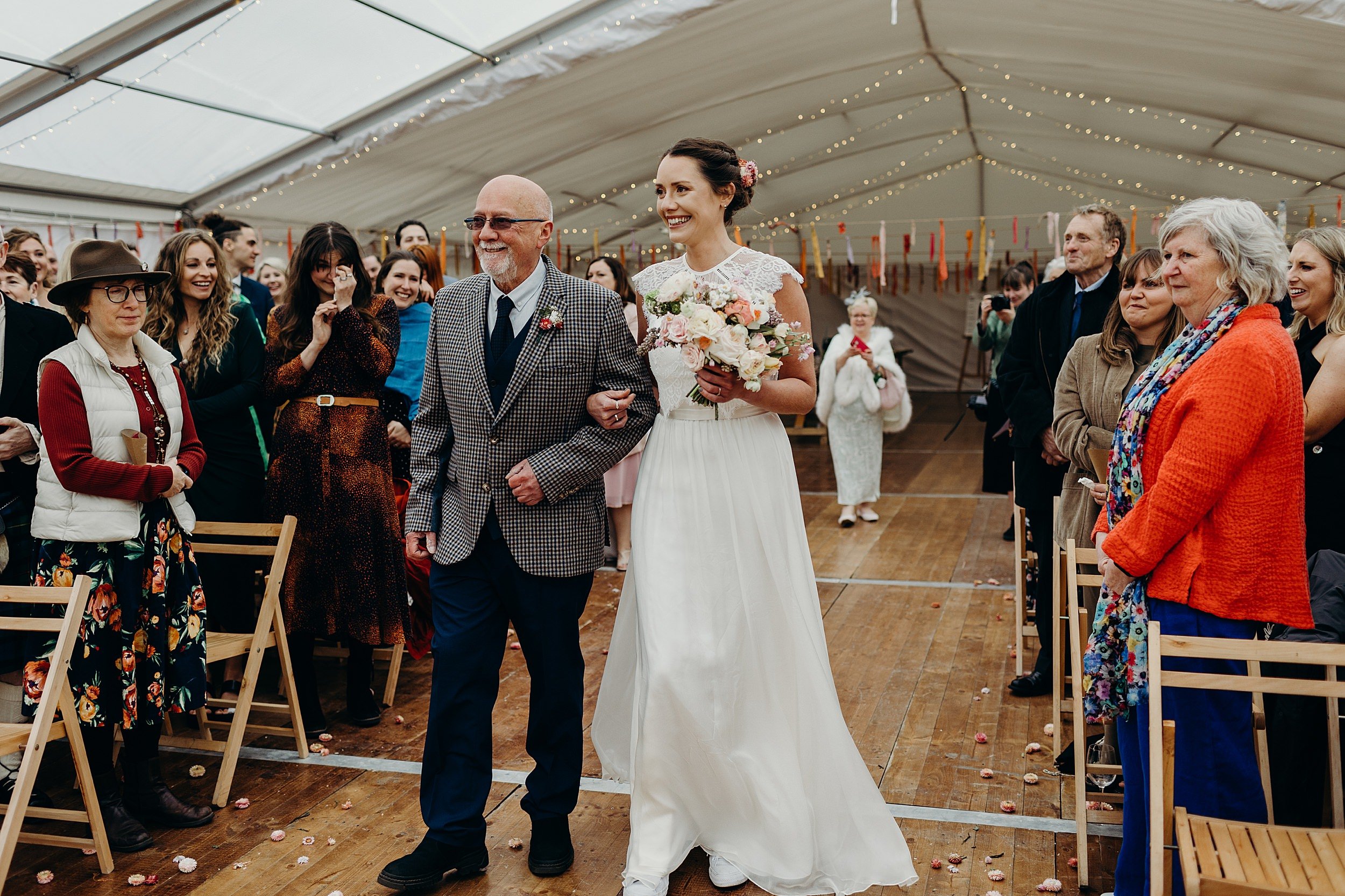 a bride arrives for her harvest moon wedding arm in arm with her father as guests stand and watch