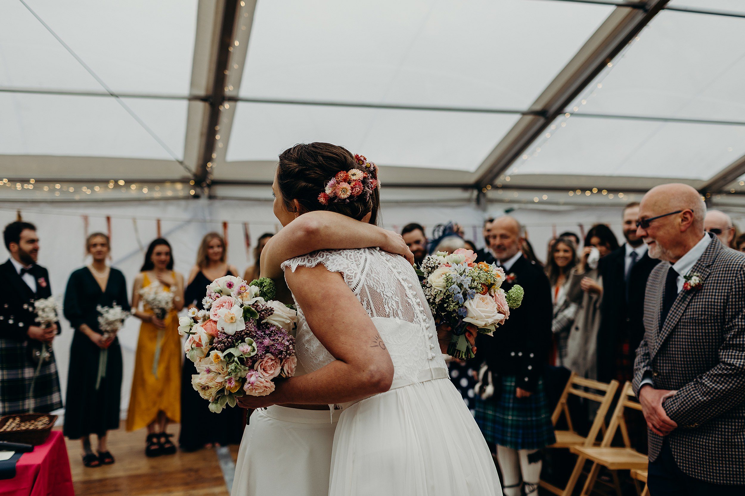 the two brides embrace as bridesmaids and guests look on before their harvest moon wedding in dunbar in scotland