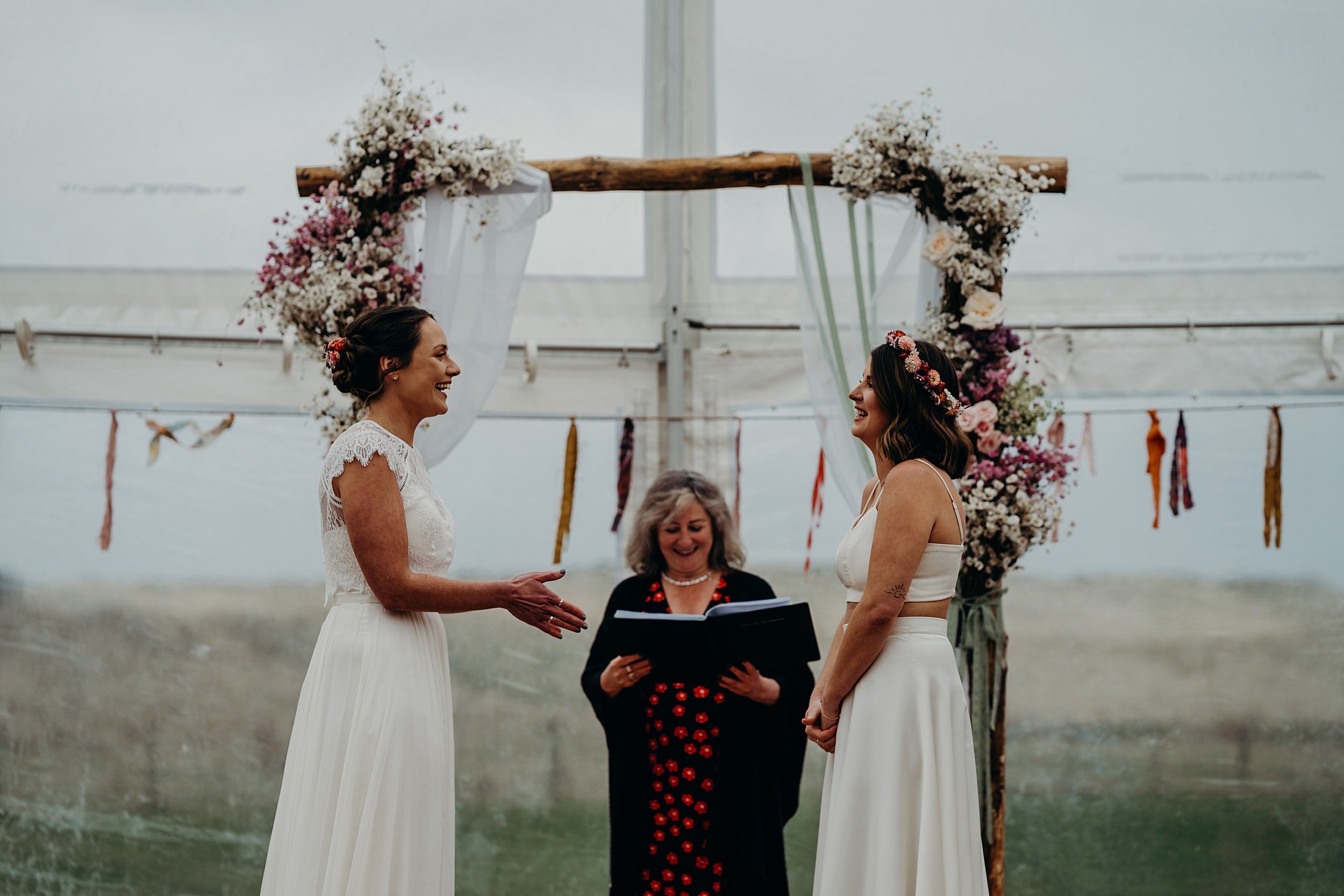 two brides stand facing each other and smiling in front of a wooden arbour inside a marquee as the celebrant conducts their harvest moon wedding in dunbar scotland