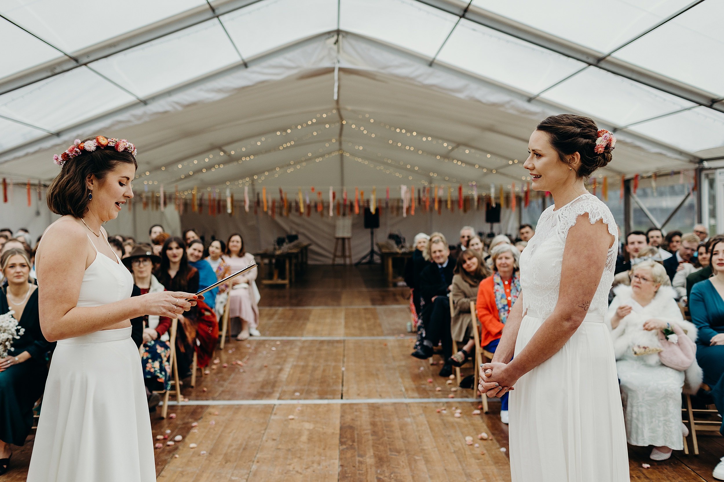 one bride reads as the other listens during their harvest moon wedding inside a white marquee as seated guests look on beneath festoon lights and ribbon garlands