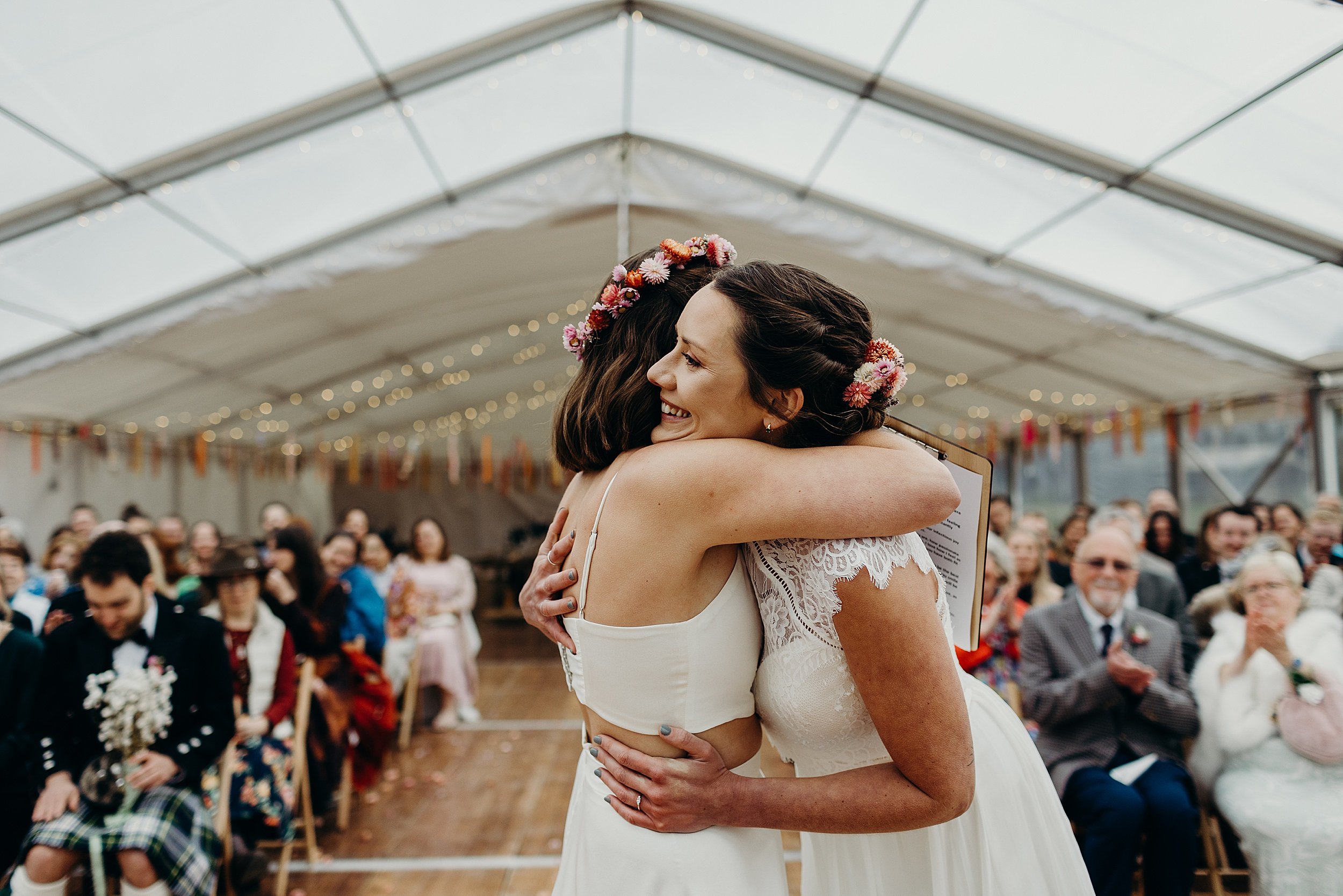 two brides embrace during their harvest moon wedding inside a white marquee as seated guests  look on beneath festoon lights and ribbon garlands