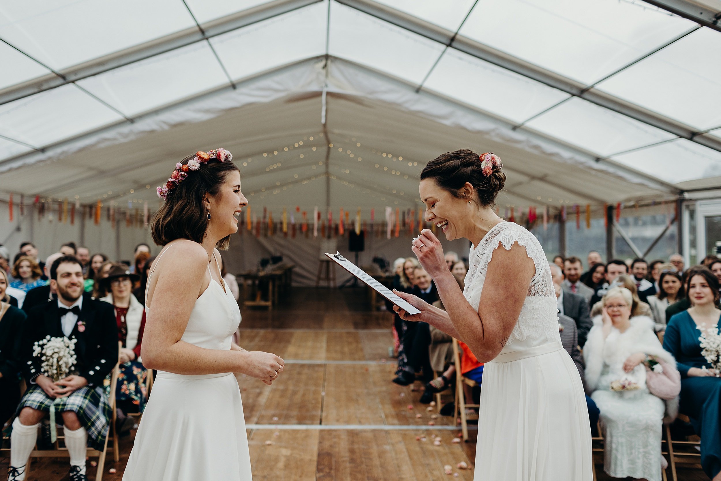 one bride reads as the other listens during their harvest moon wedding inside a white marquee as seated guests look on beneath festoon lights and ribbon garlands