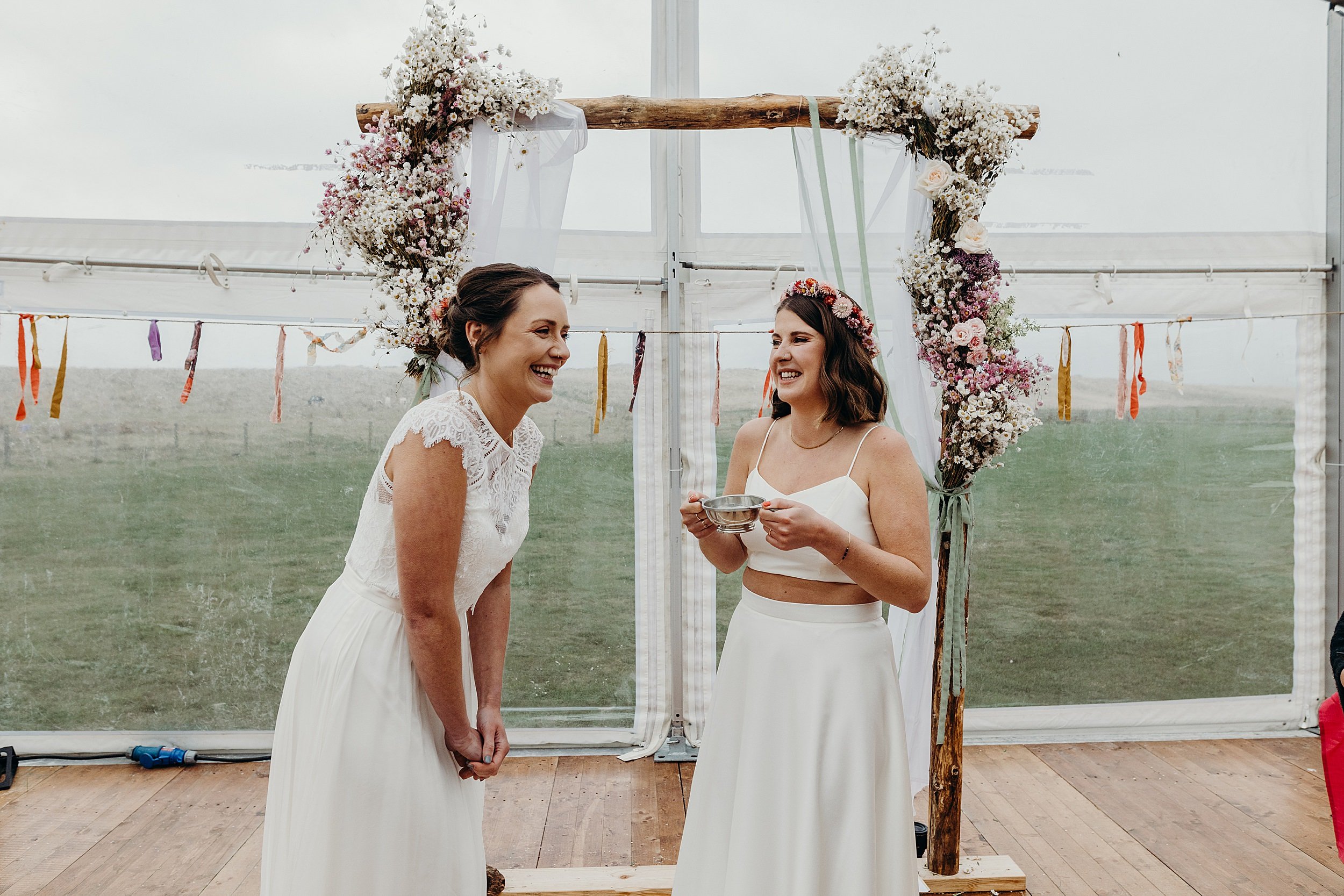 standing on front of a wooden arbour inside a white marquee one bride holds a quaich while the other smiles
