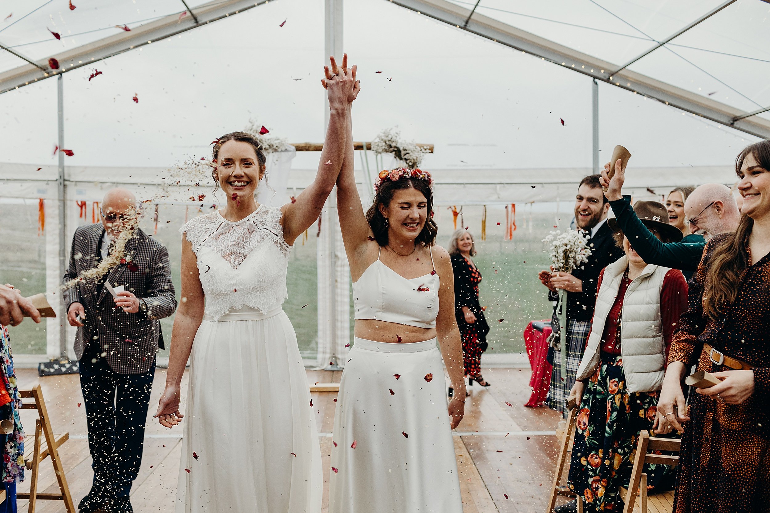 inside a white marquee guests throw confetti as the two brides walk down the aisle following their harvest moon wedding in dunbar east lothian scotland