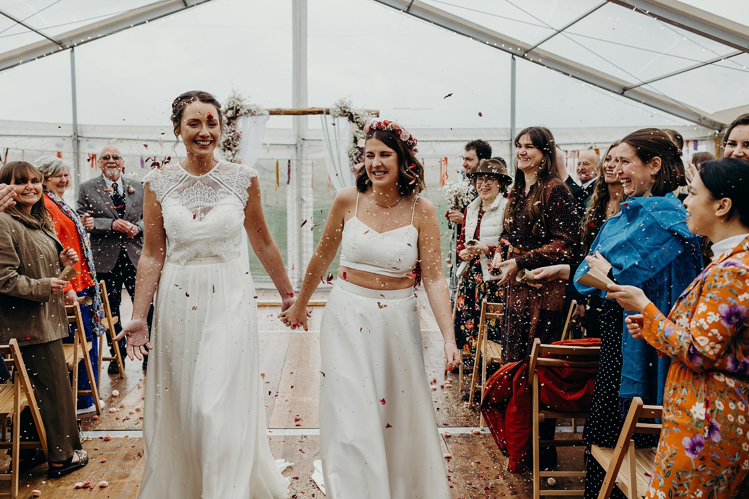 inside a white marquee guests throw confetti as the two brides walk down the aisle following their harvest moon wedding in dunbar east lothian scotland