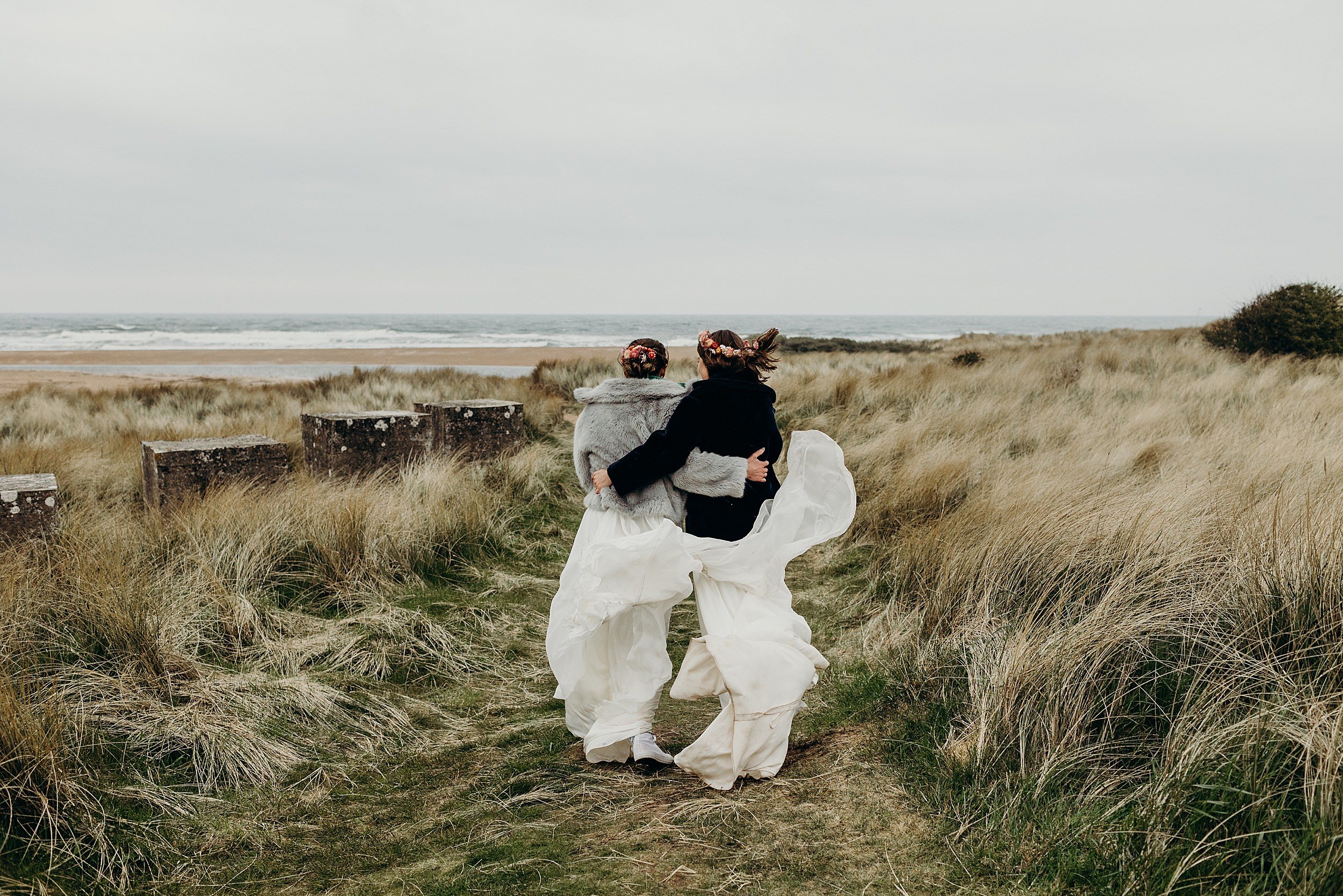 two brides wearing coats over their wedding gowns walk through sand dunes towards the sea following their harvest moon wedding in east lothian in scotland