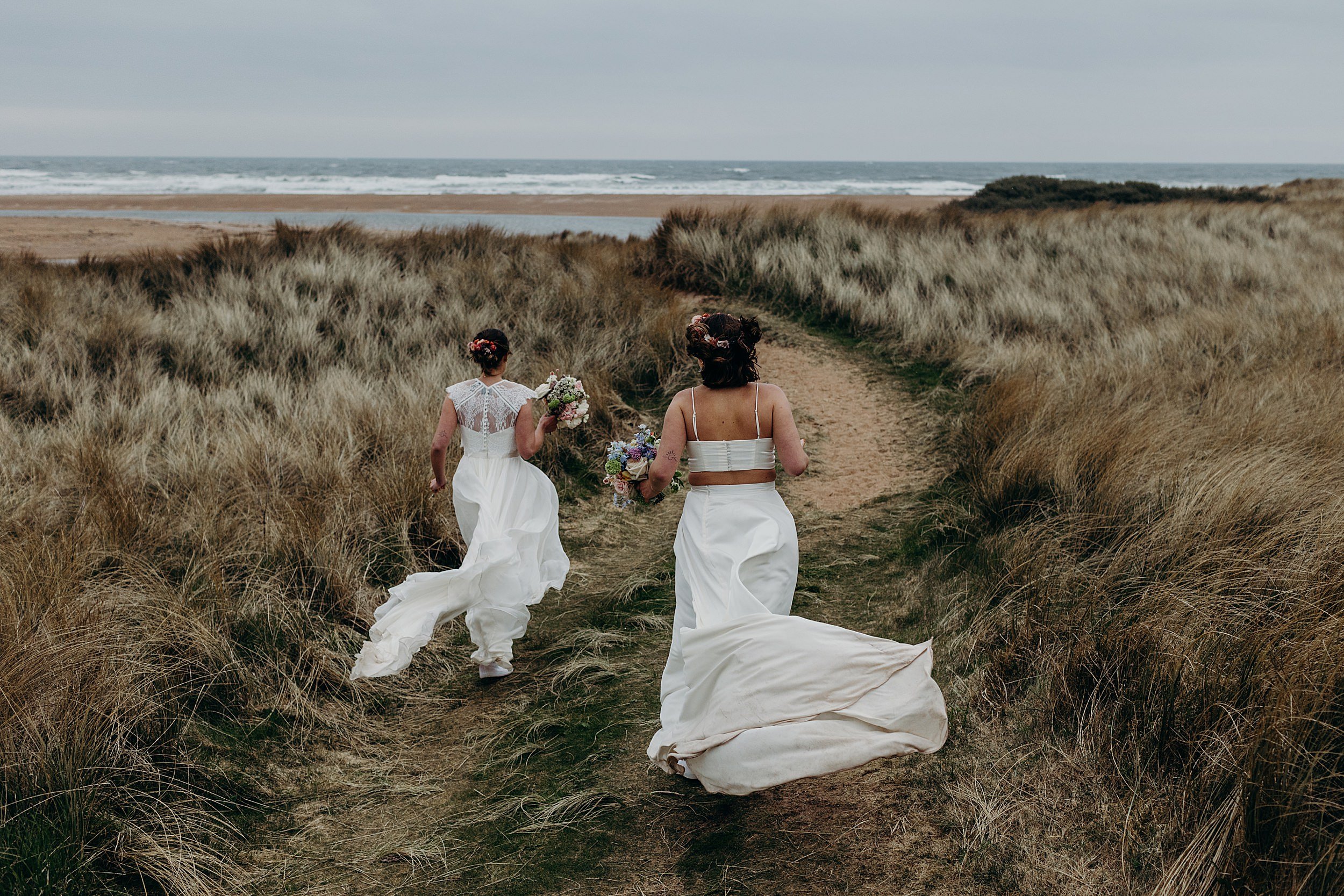 two brides wearing white dresses and carrying bouquets walk through sand dunes towards the sea following their harvest moon wedding in east lothian in scotland