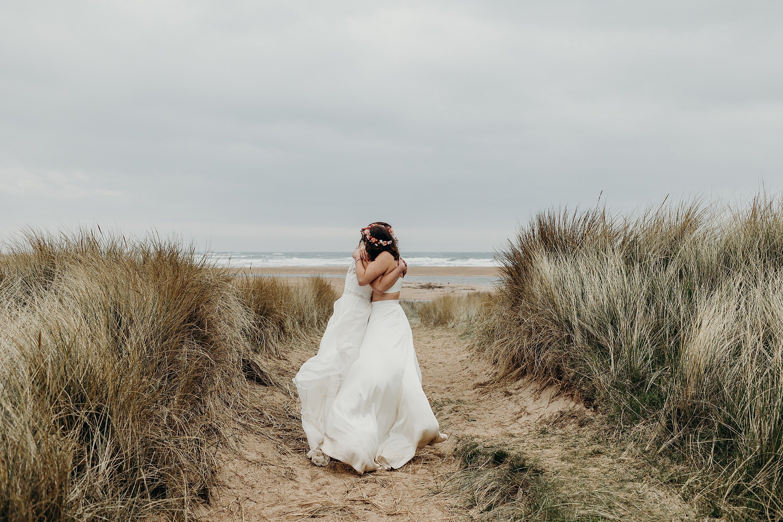 two brides wearing white dresses embrace in the sand dunes with the sea and beach visible in the distance following their harvest moon wedding in dunbar scotland