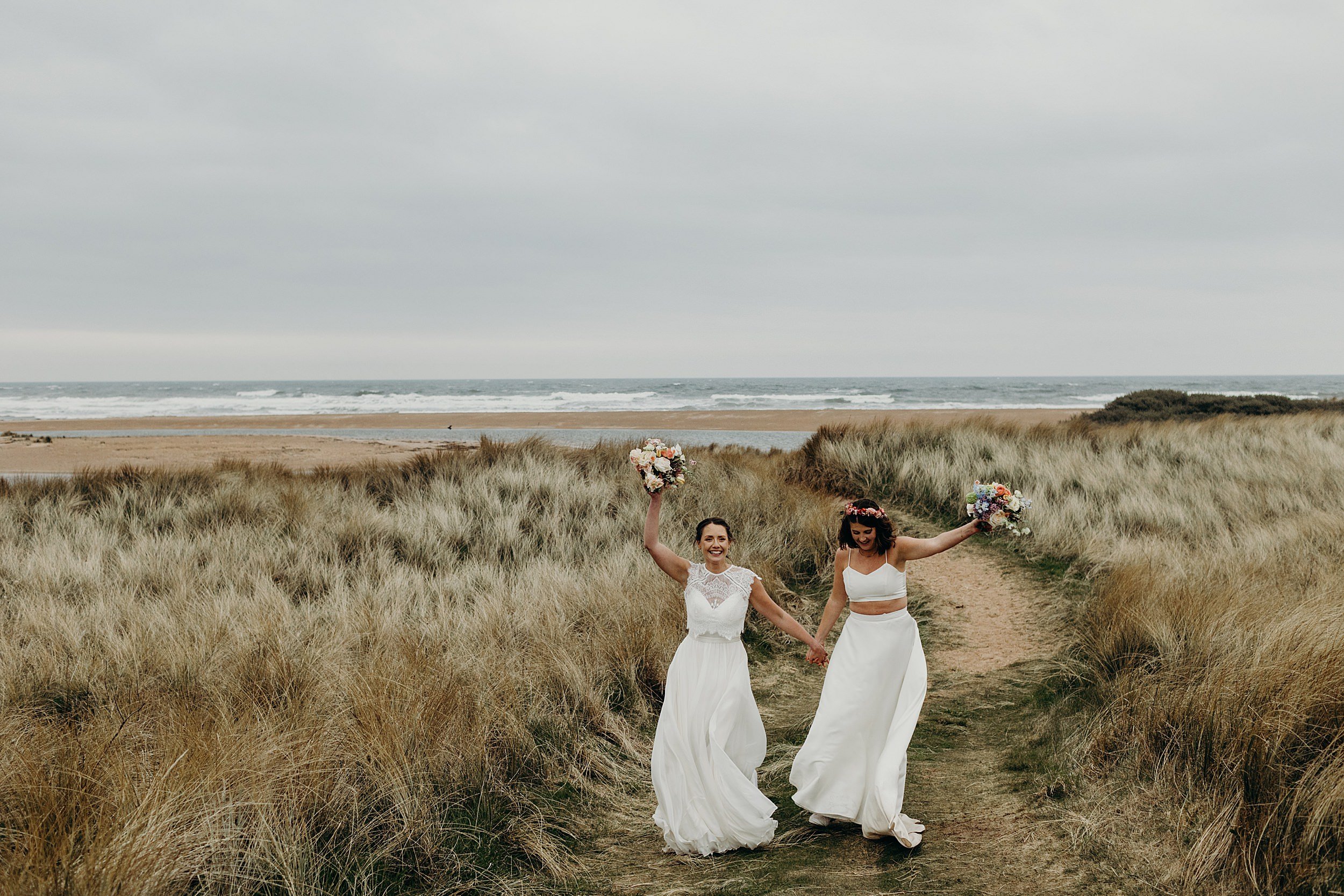 two brides wearing white hold hands and hold their bouquets above their heads as they walk away from the sea and beach following their harvest moon wedding