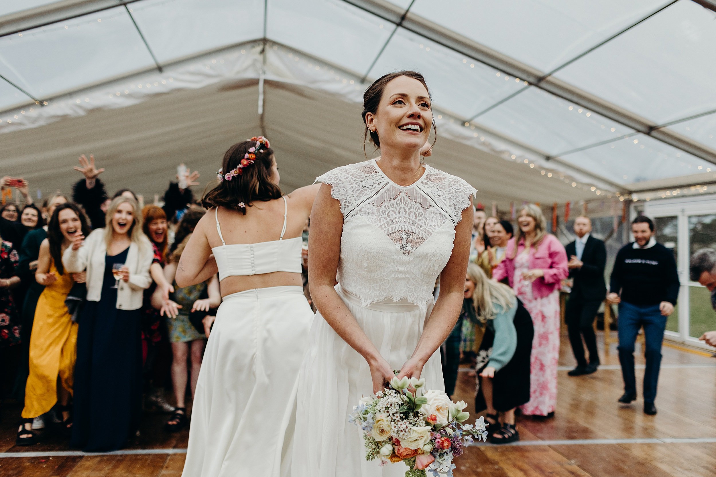 a bride prepares to toss her bouquet into the crowd of guests inside a white marquee at harvest moon wedding venue in East Lothian in Scotland