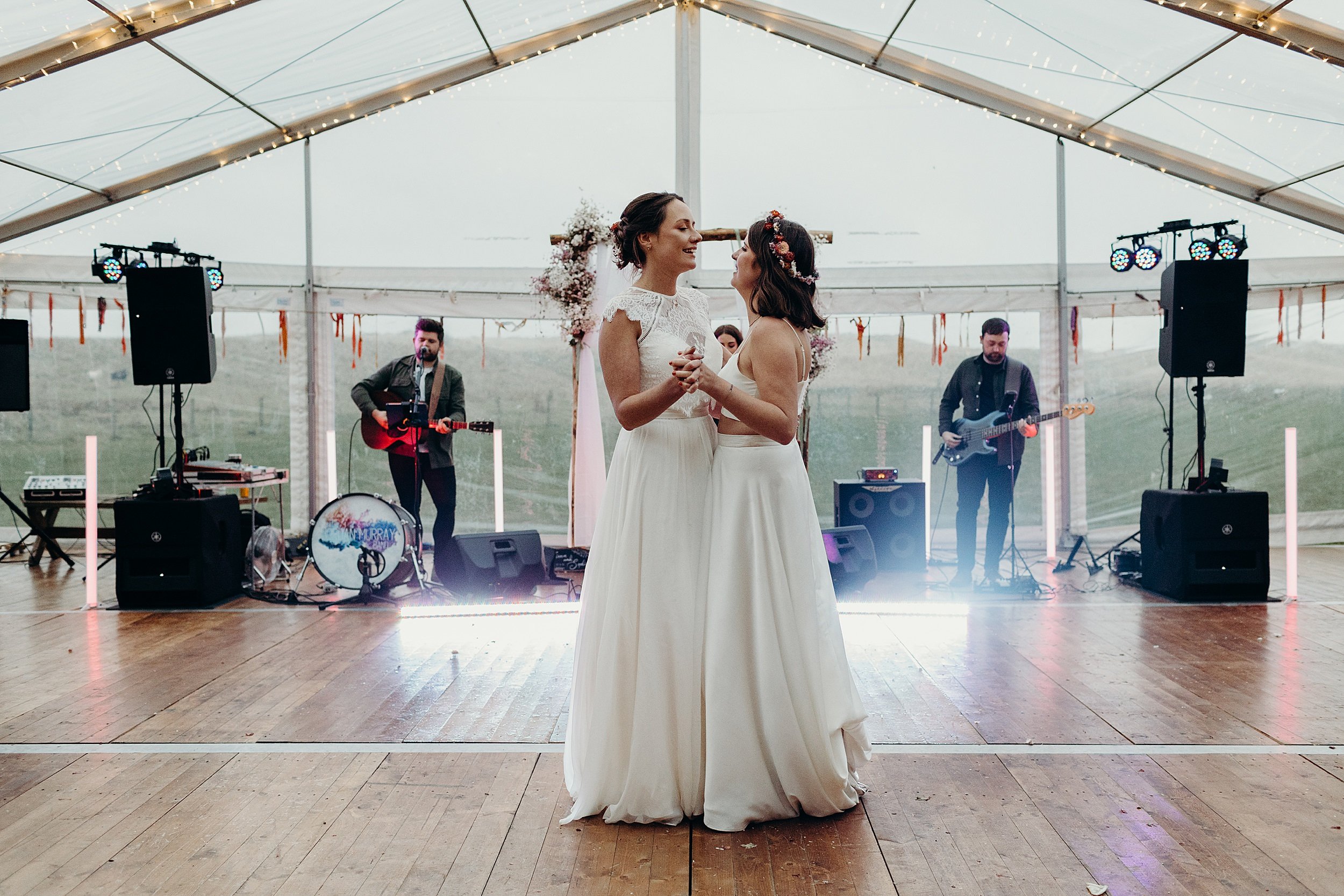the two brides dance their first dance in a white marquee at harvest moon wedding venue in dunbar scotland a band is visible playing in the background