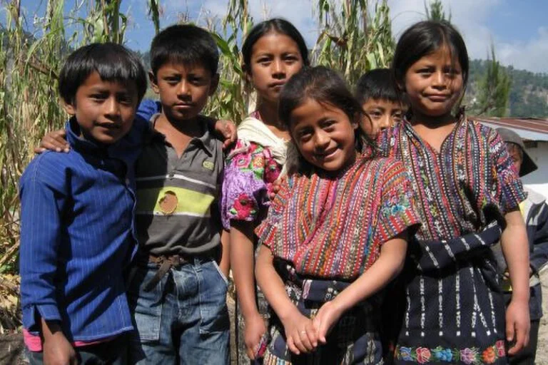 Group of five children with dark hair and brown skin, wearing traditional colorful clothing, standing outdoors in front of tall grass or crops, smiling and looking at the camera.
