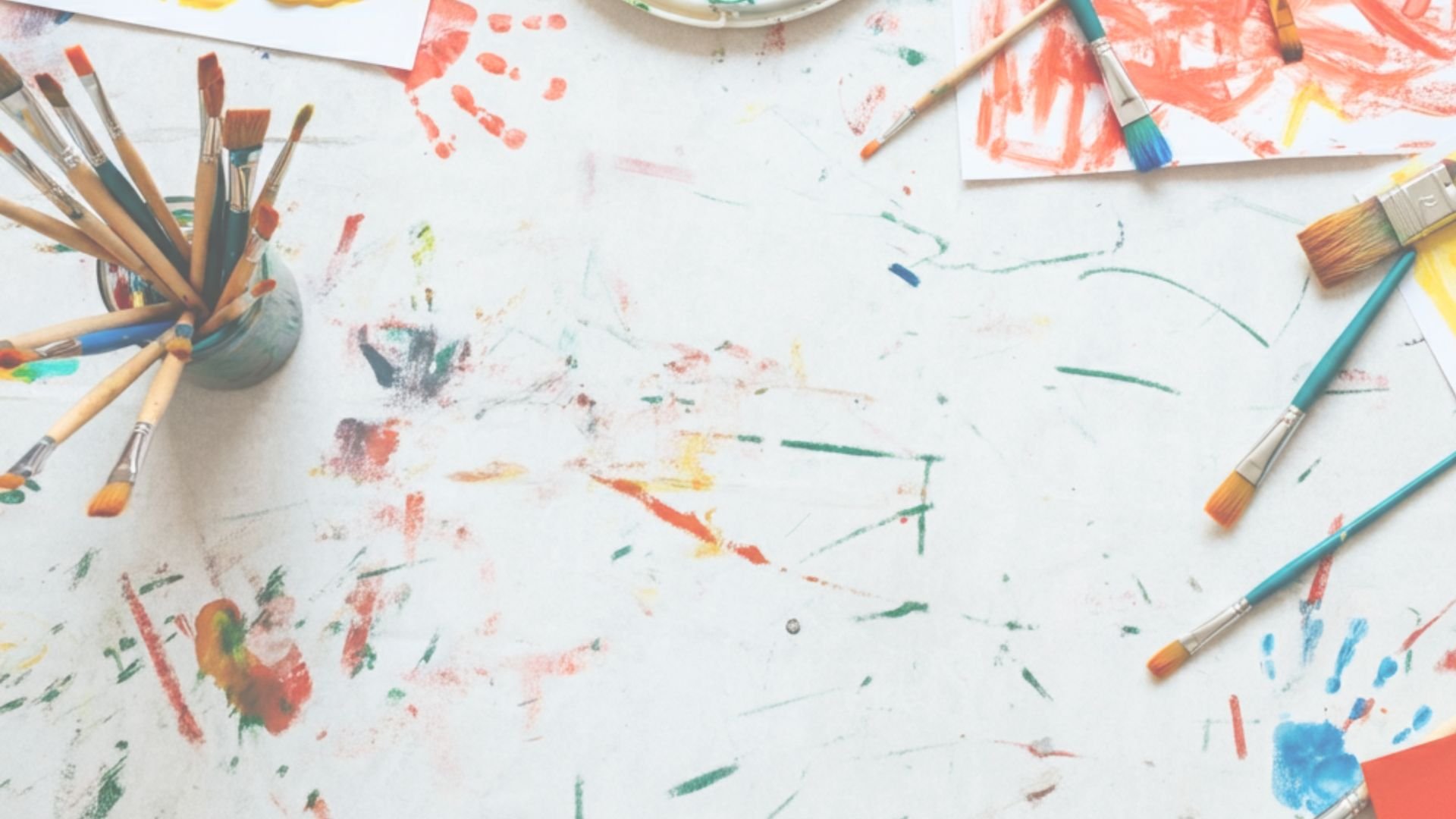 A young girl with long blonde hair painting with watercolors at a desk covered with art supplies.
