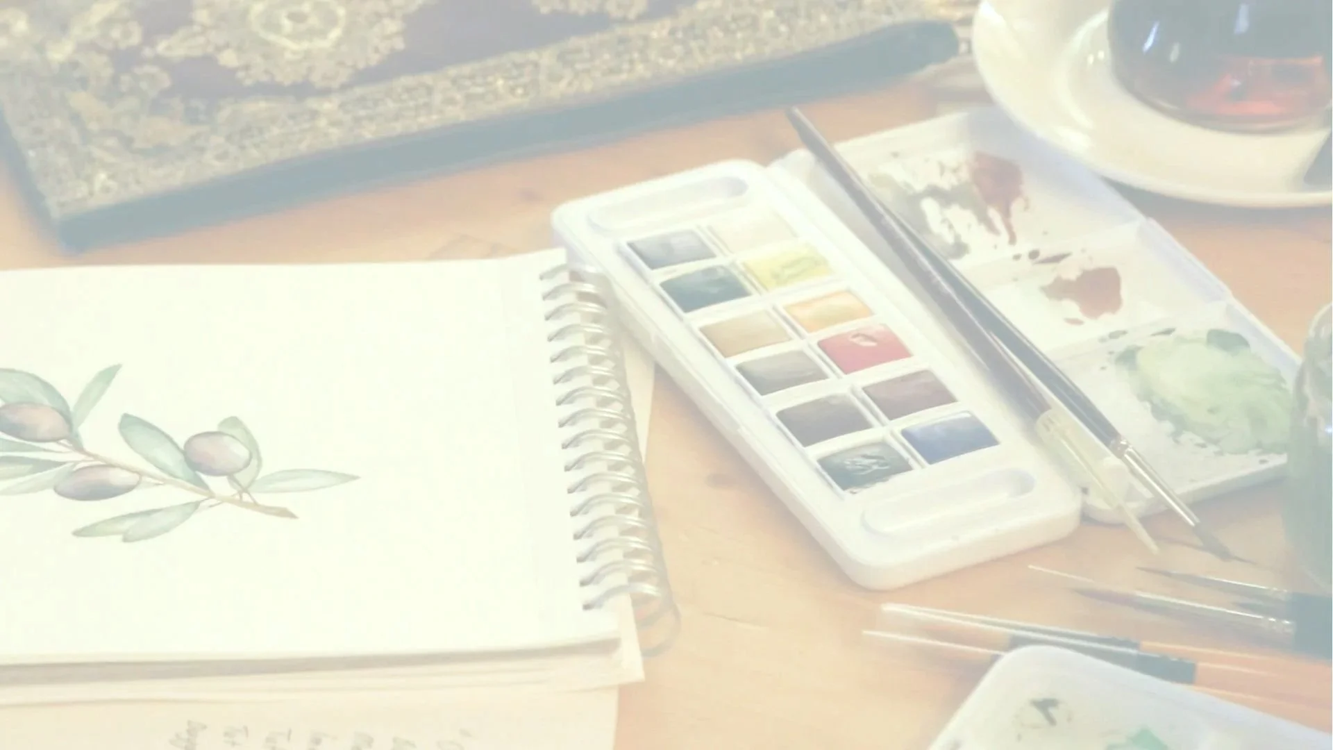 Person painting a watercolor illustration of oranges and green leaves on white paper, with a watercolor palette nearby.
