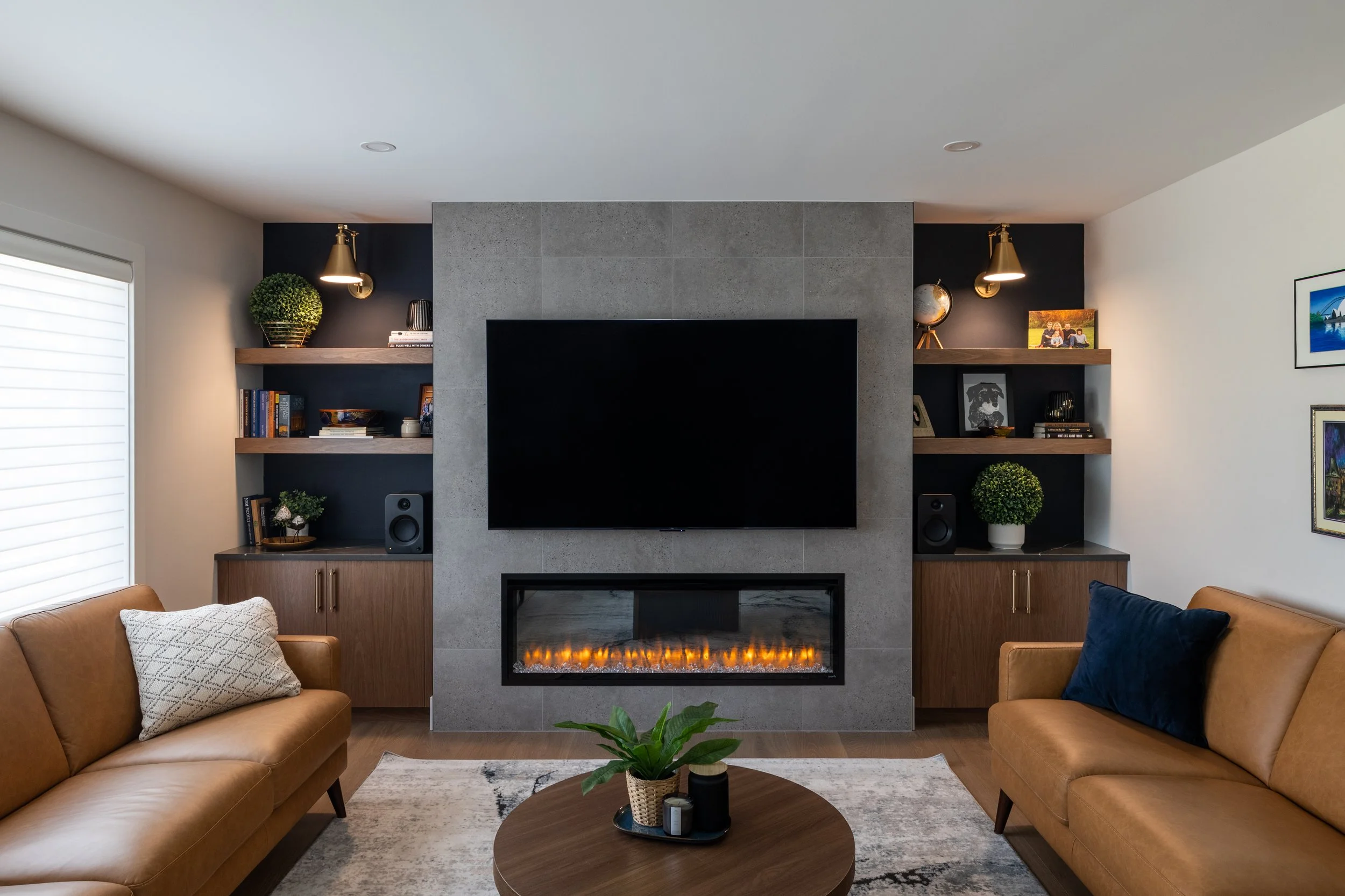 A modern living room with two leather sofas facing each other, a coffee table with decor, and a fireplace with a TV mounted above it. The room features built-in shelves with books and decorations, and has large windows.
