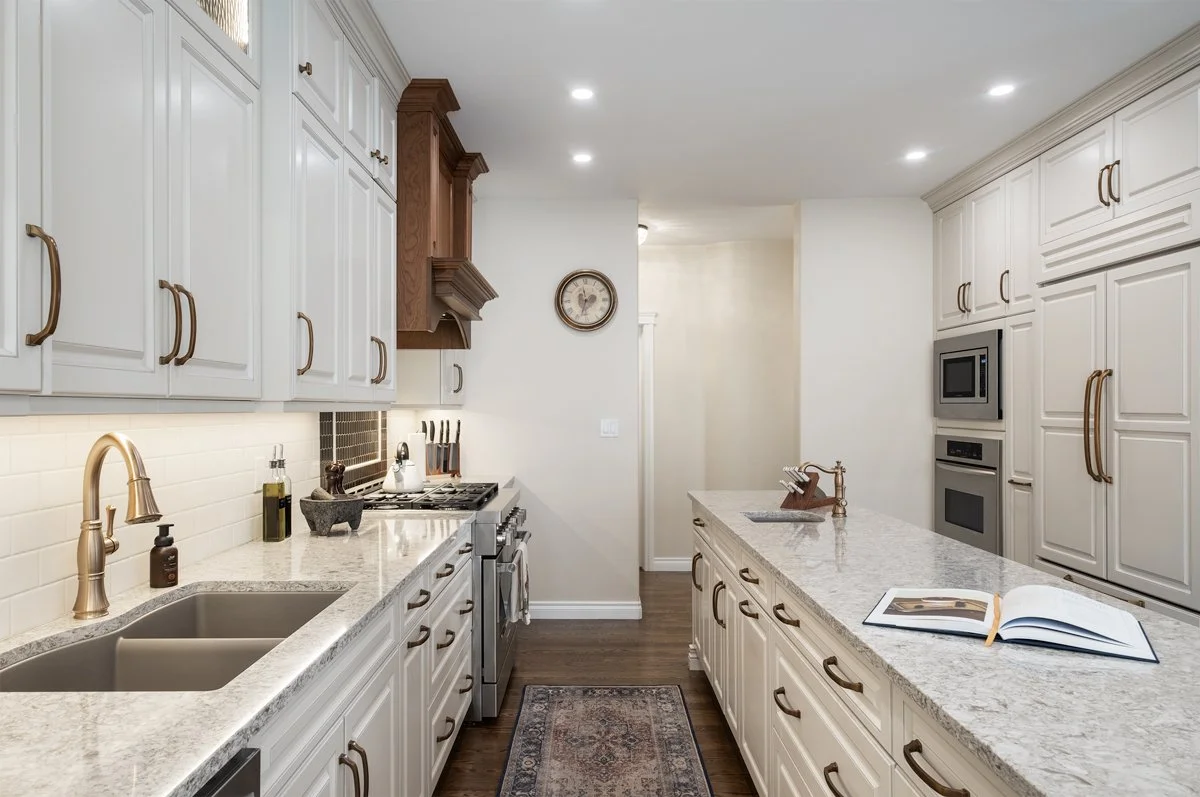 A spacious kitchen with white cabinets, a large island with a granite countertop, and stainless steel appliances. There is a double sink with a gold faucet, a stove with a hood, and a built-in microwave and oven. The floor is wooden, and there is a small rug in front of the sink. A clock is mounted on the wall above the counter.