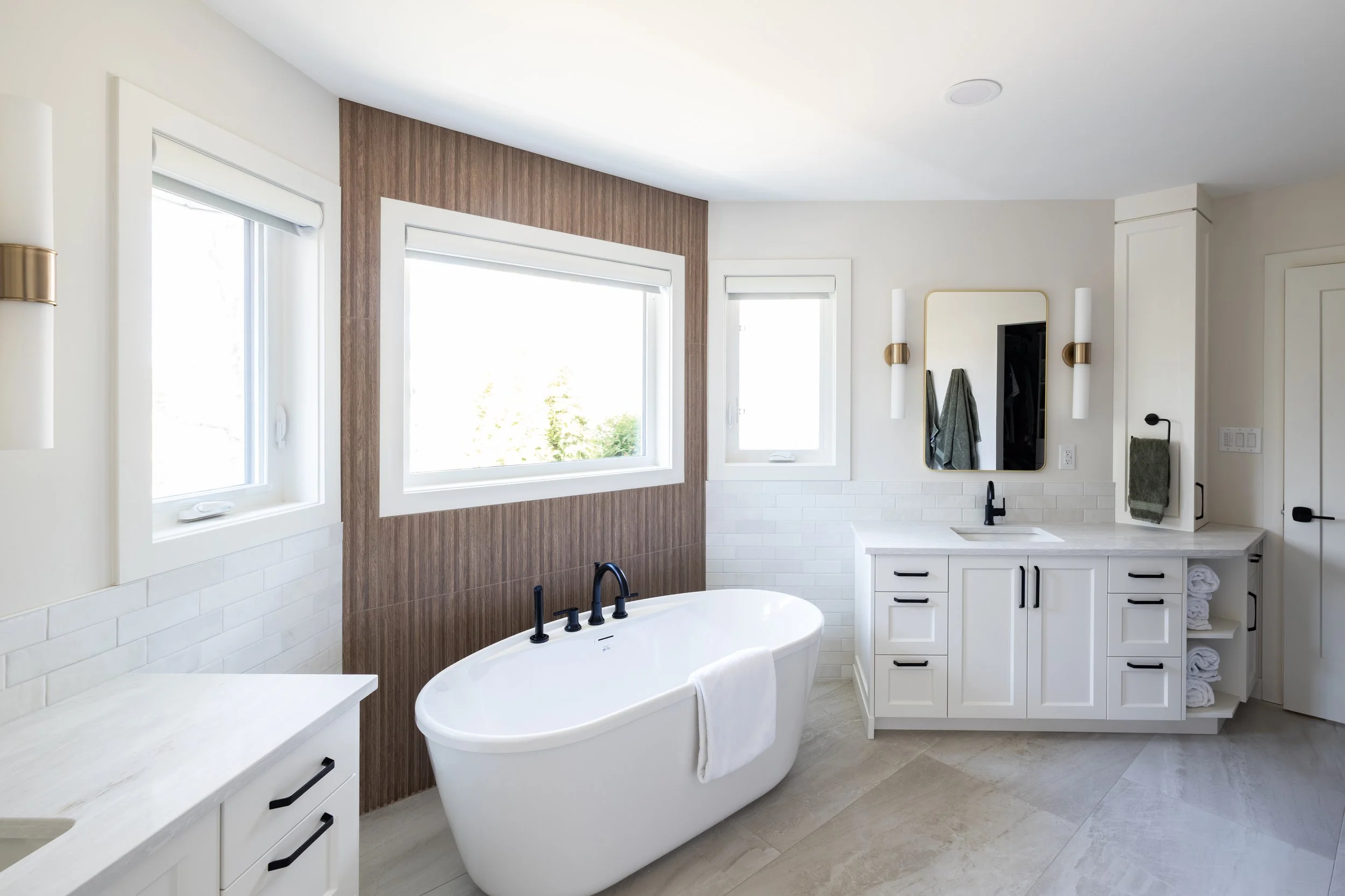 Modern bathroom with a standalone bathtub, white vanity with black handles, a mirror, and windows with white blinds.