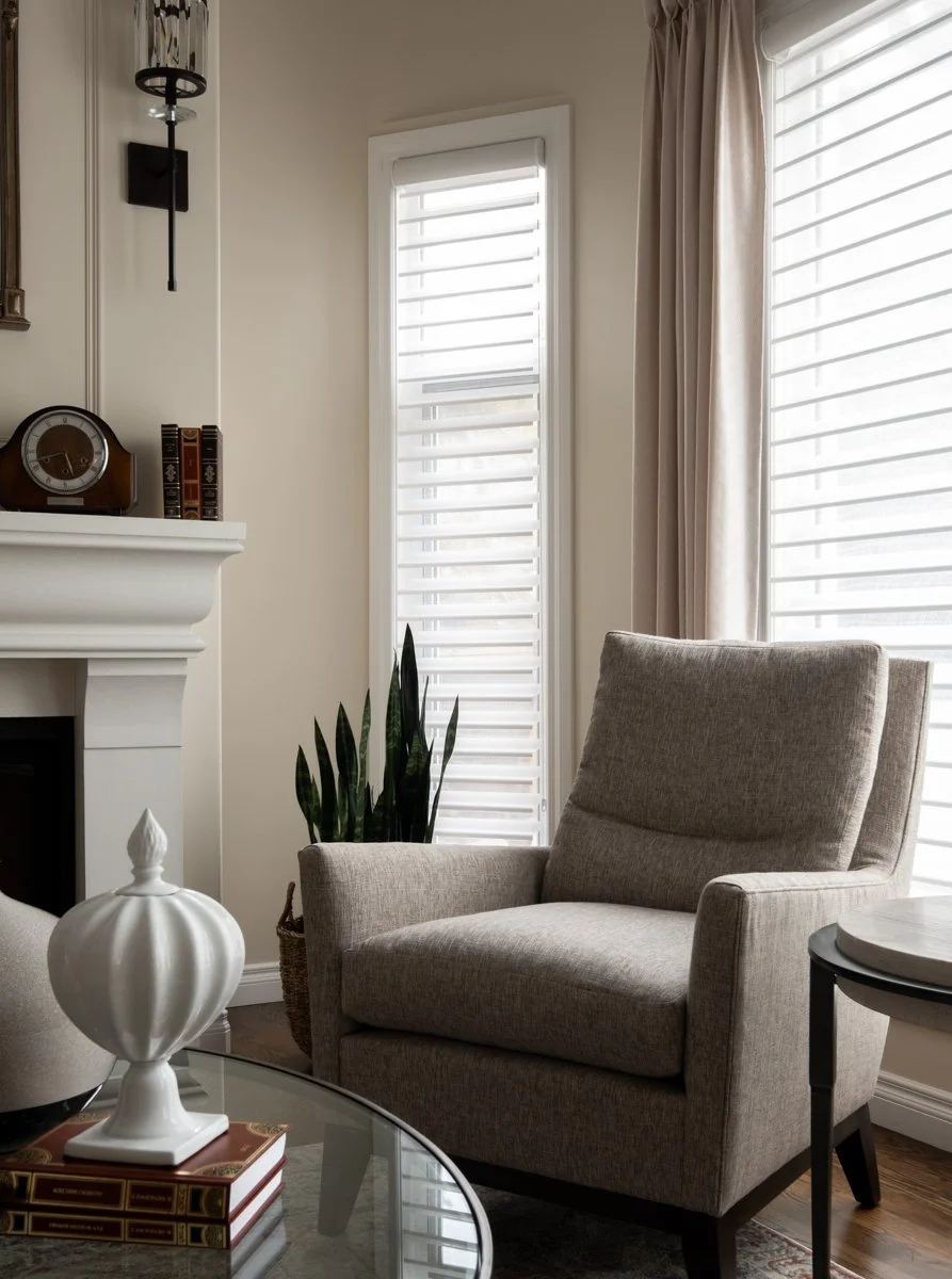 Living room corner with a beige armchair, potted plant, glass-topped coffee table, and window with white blinds and beige curtains.