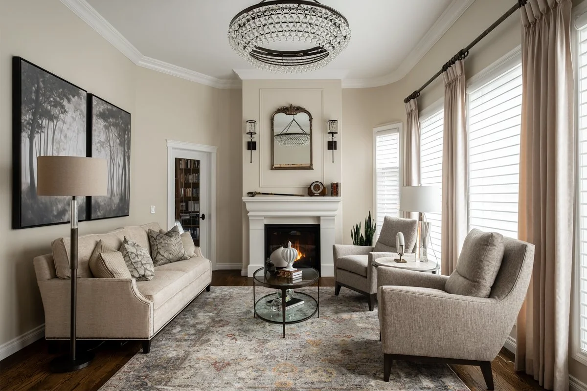 Living room with beige sofa, two beige armchairs, a round glass coffee table, fireplace, wall-mounted mirror, large window with beige curtains, wall art, and chandelier ceiling light.