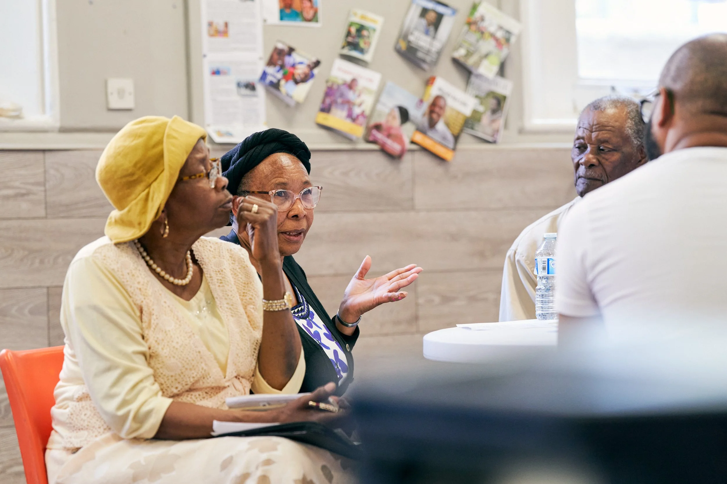 Four people are seated and engaged in a discussion in a room. The woman on the left is wearing a yellow headwrap and glasses, listening attentively while holding some papers. The woman next to her is wearing a black headwrap and glasses, gesturing wi