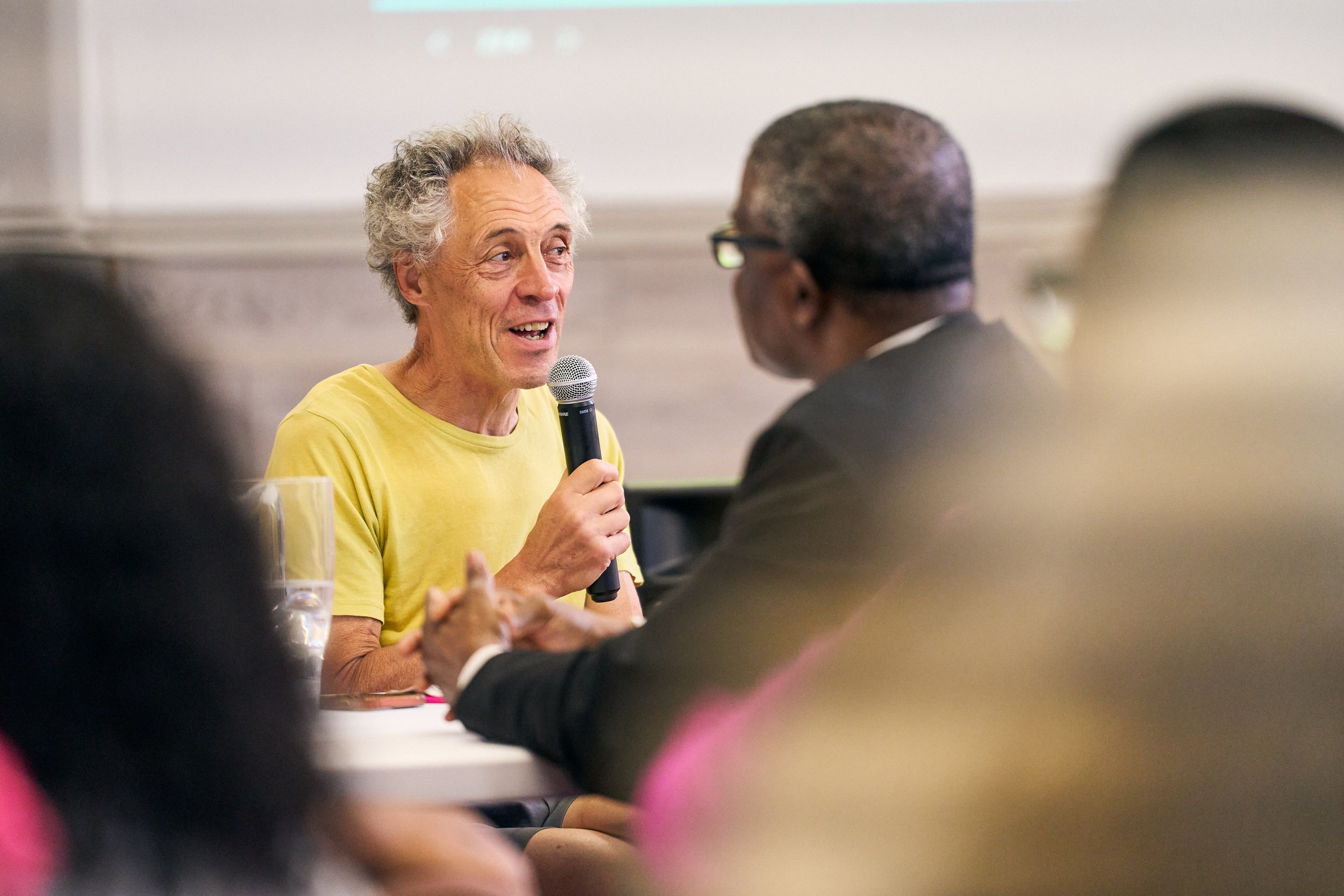 An older man with curly gray hair wearing a yellow shirt, speaking into a microphone at a discussion or presentation, while a person with glasses, dark hair, and wearing a suit listens attentively.